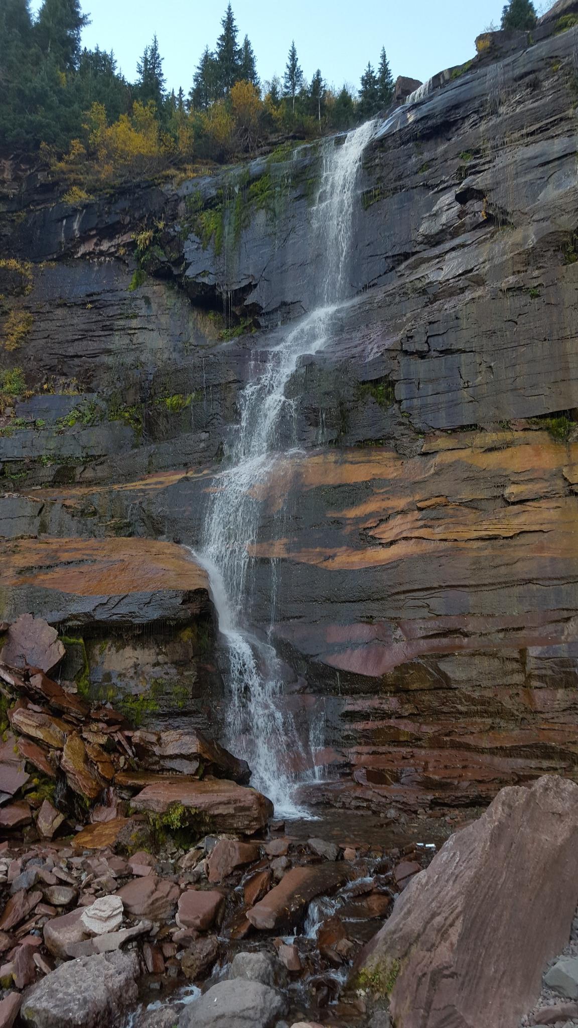 A tall waterfall cascades down a layered rock cliff with autumn trees above.