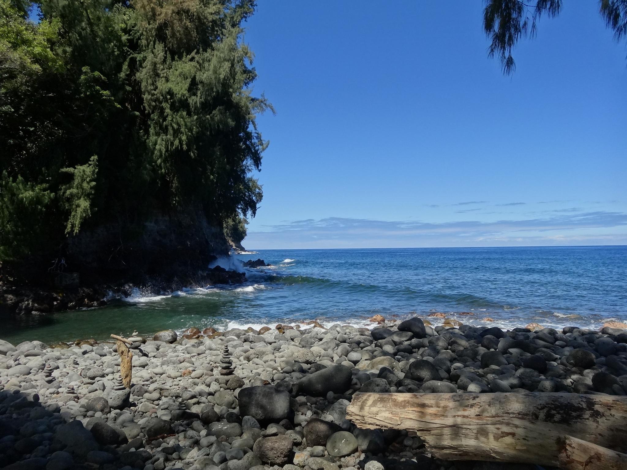 Sunny rocky beach with ocean waves, a lush green cliff, and stacked rocks.