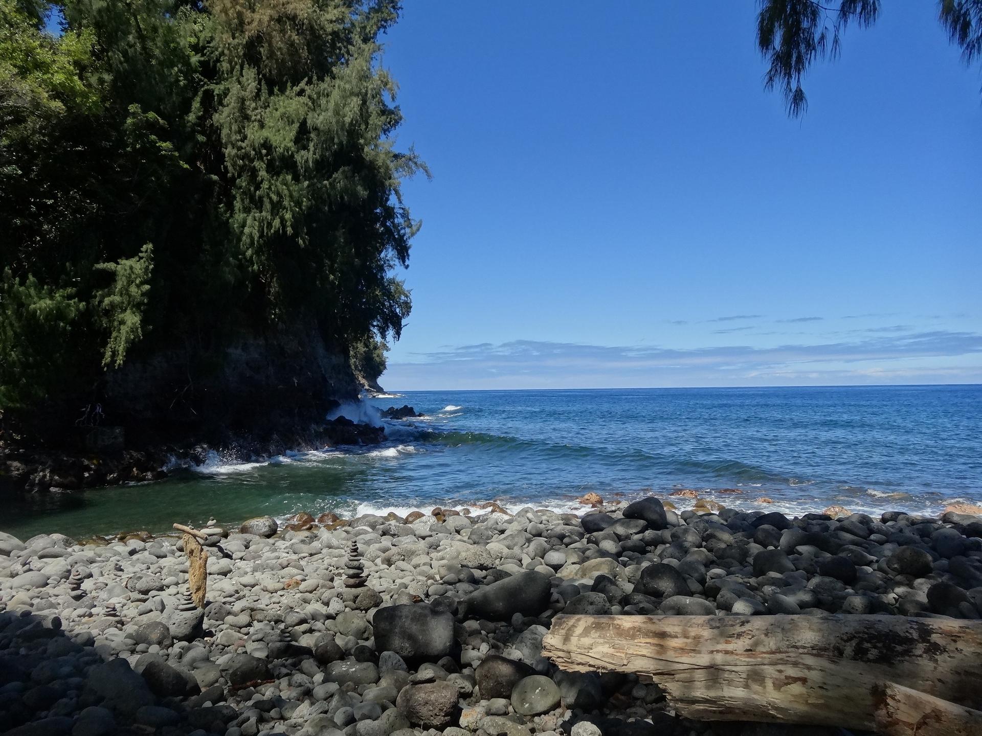 Sunny rocky beach with ocean waves, a lush green cliff, and stacked rocks.