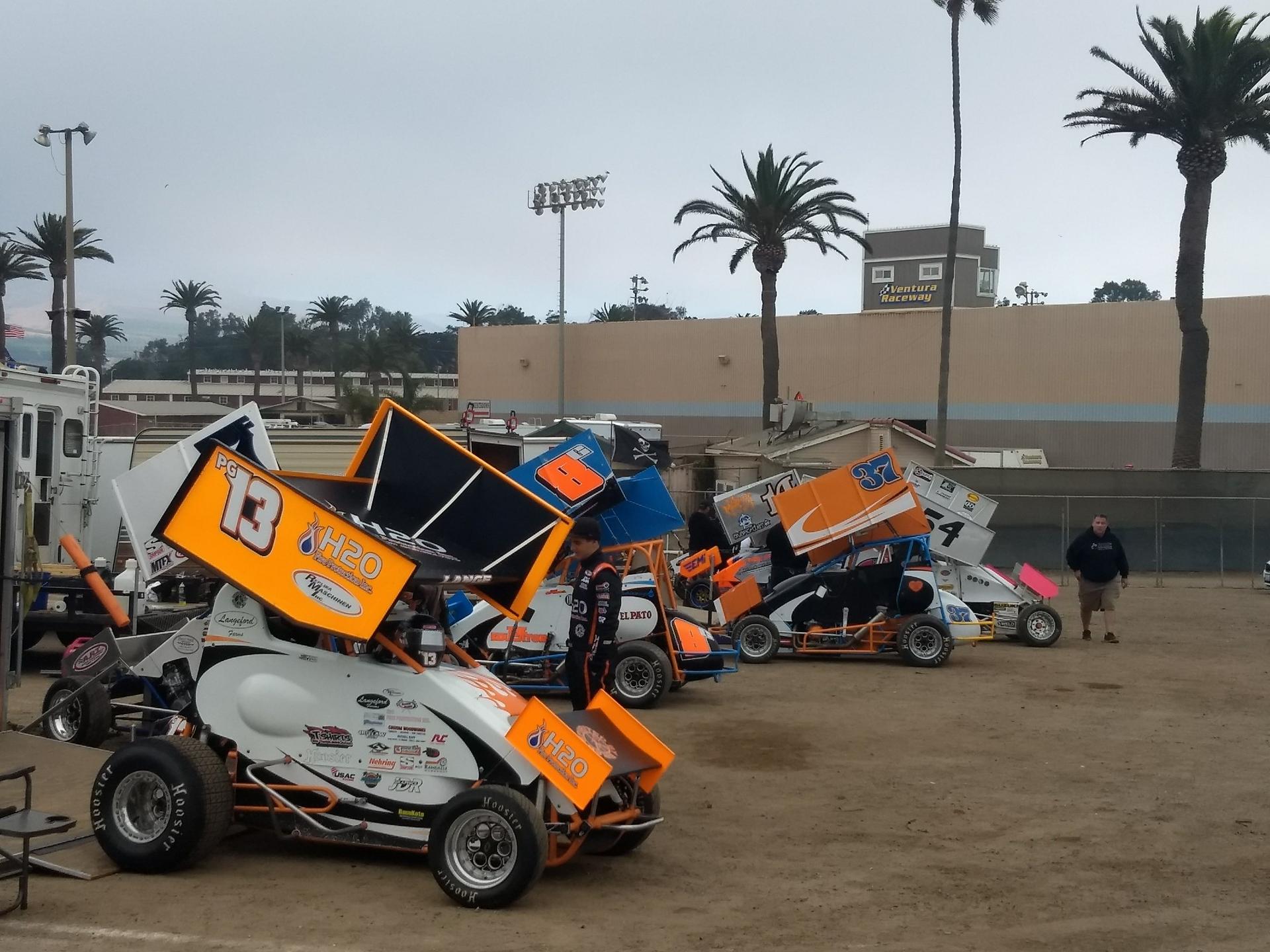 Several sprint cars parked on a dirt track with palm trees and buildings.