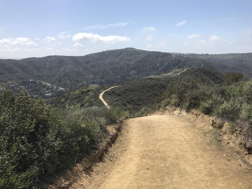 A wide dirt path winds through green hills under a partly cloudy sky.