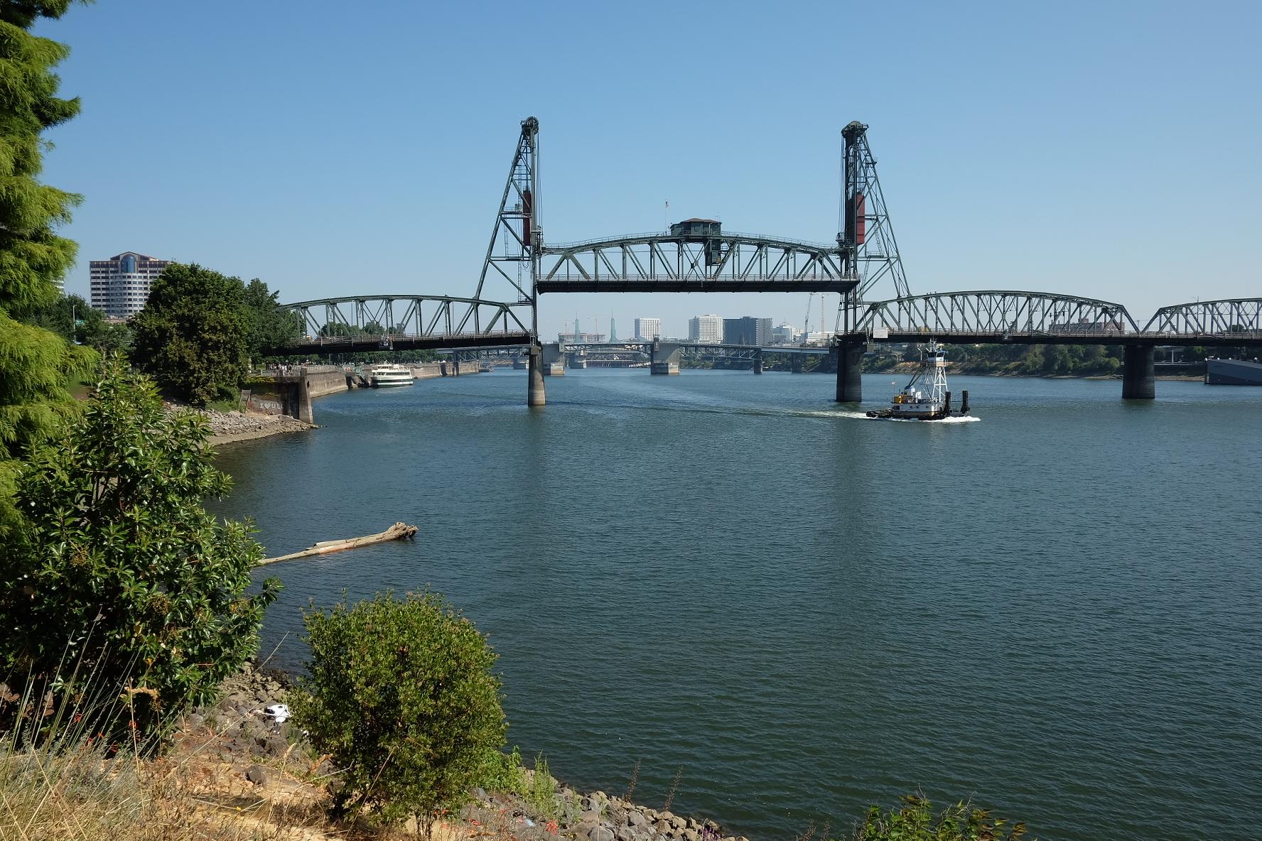 A large metal bridge spans a river with a tugboat passing underneath.
