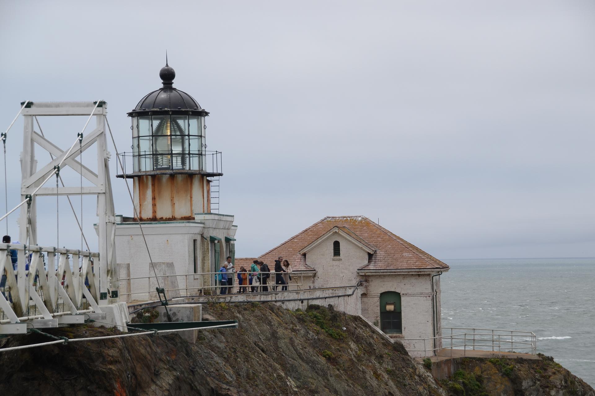 Historic lighthouse and building on a cliff edge with people, under a gray sky.