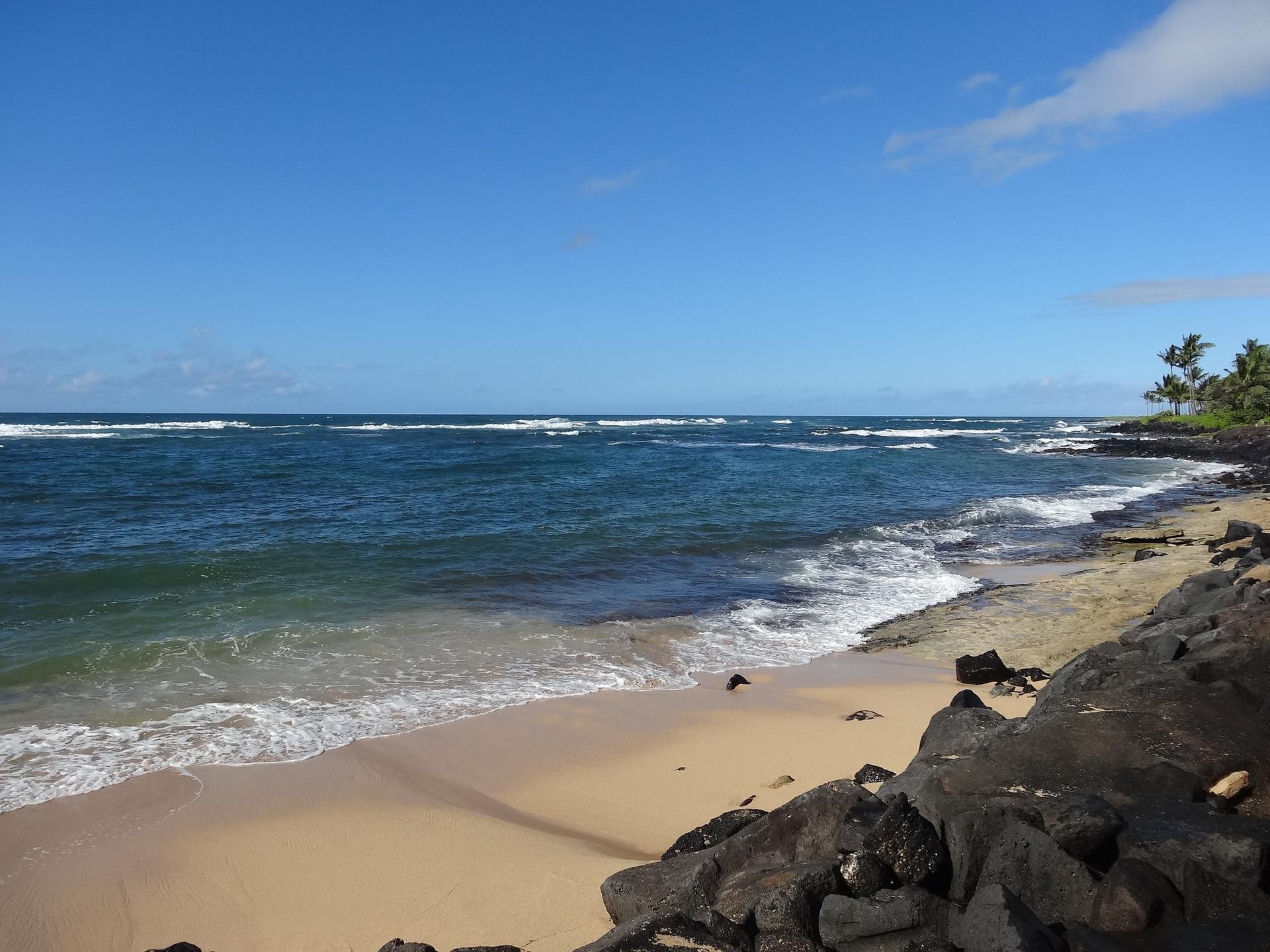 Sunny sandy beach with gentle waves and dark rocks in the foreground.