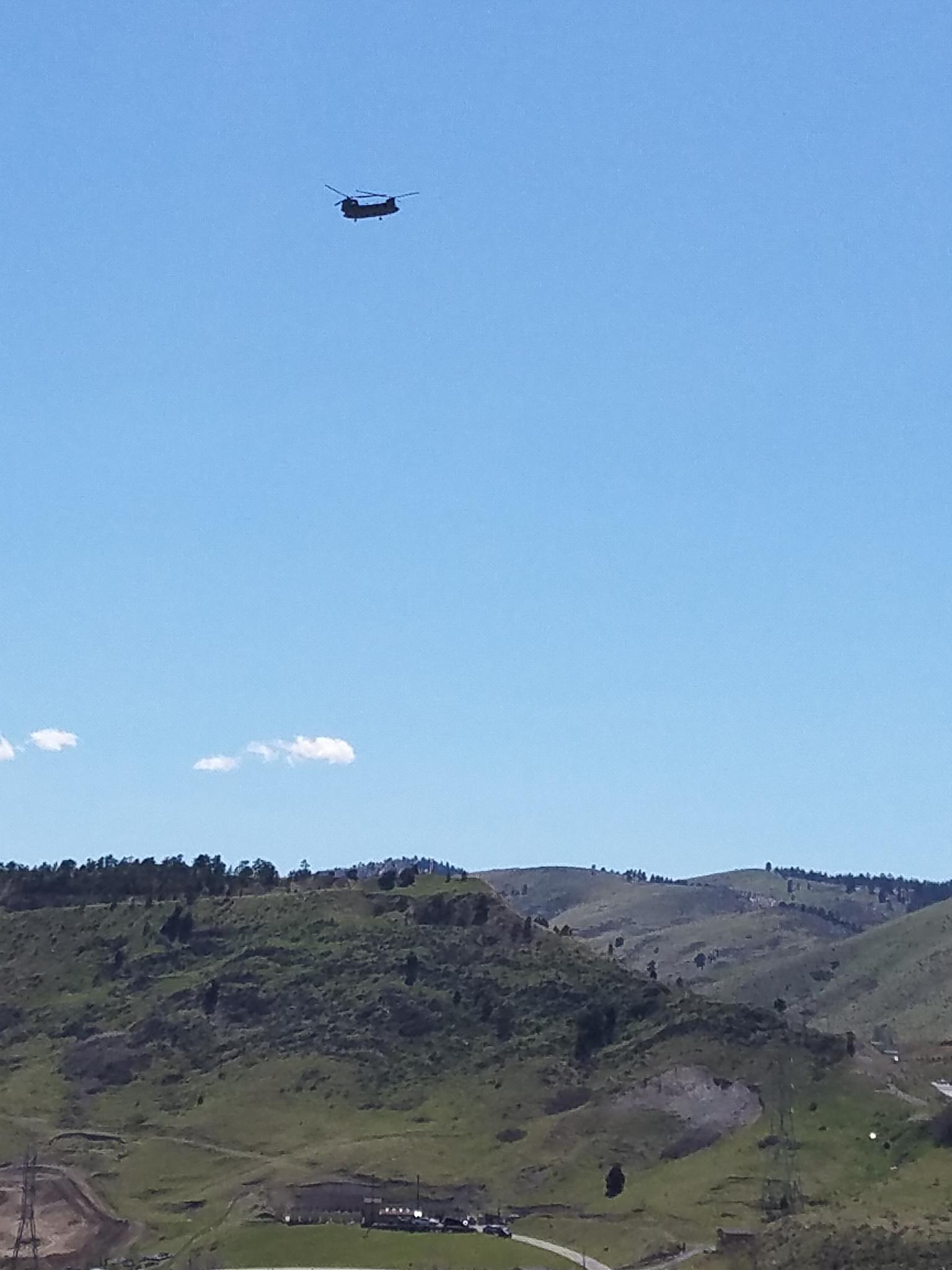 A helicopter flies high above green rolling hills on a clear day.