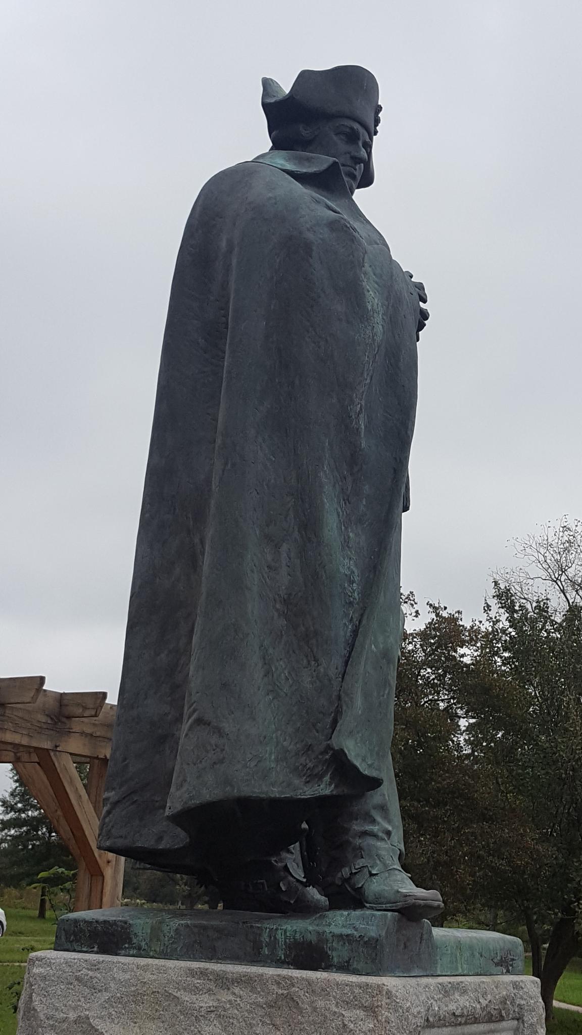 Bronze statue of a man in a cape and hat, seen from below, cloudy sky.