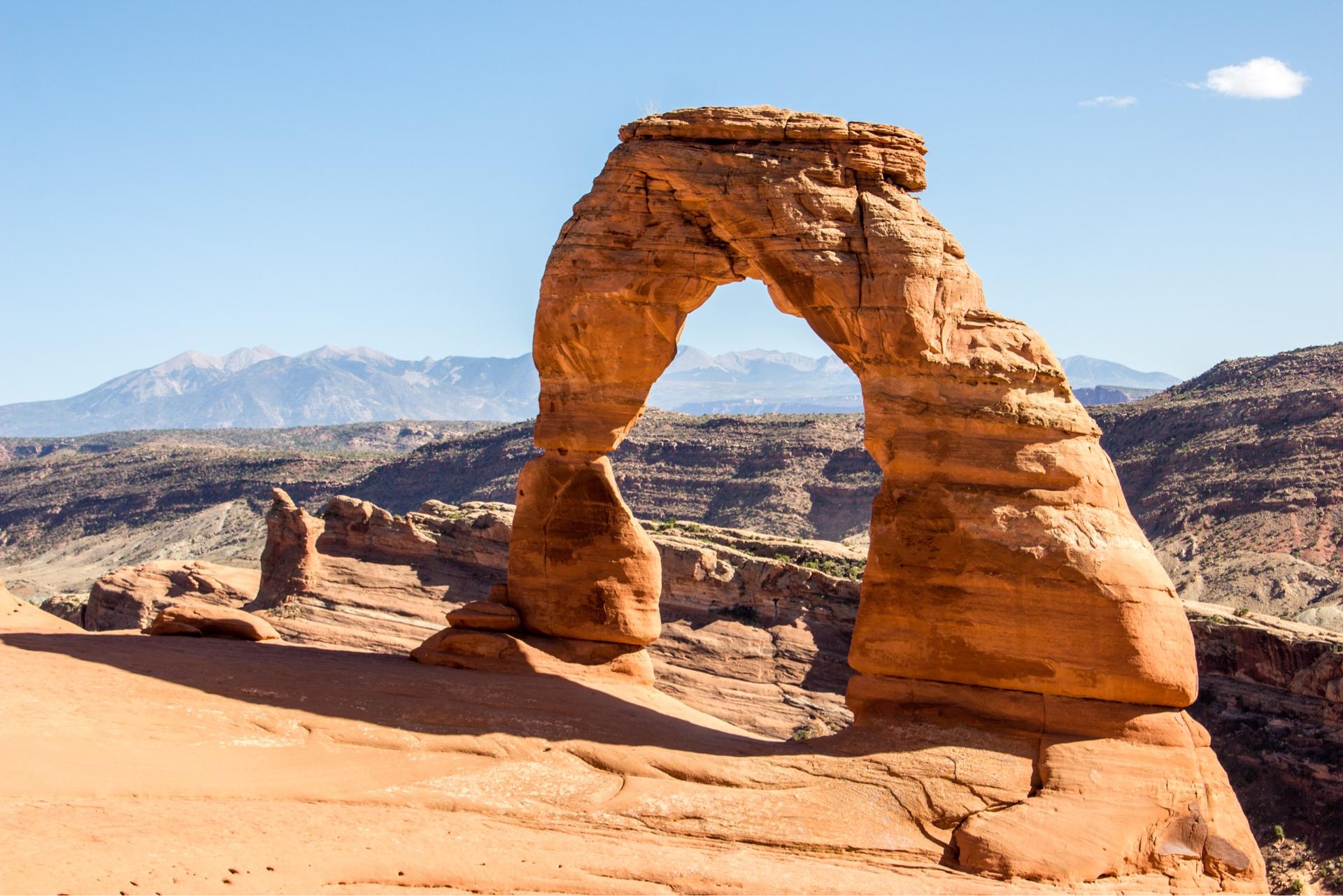 A large natural arch in a sunny desert landscape with distant mountains.