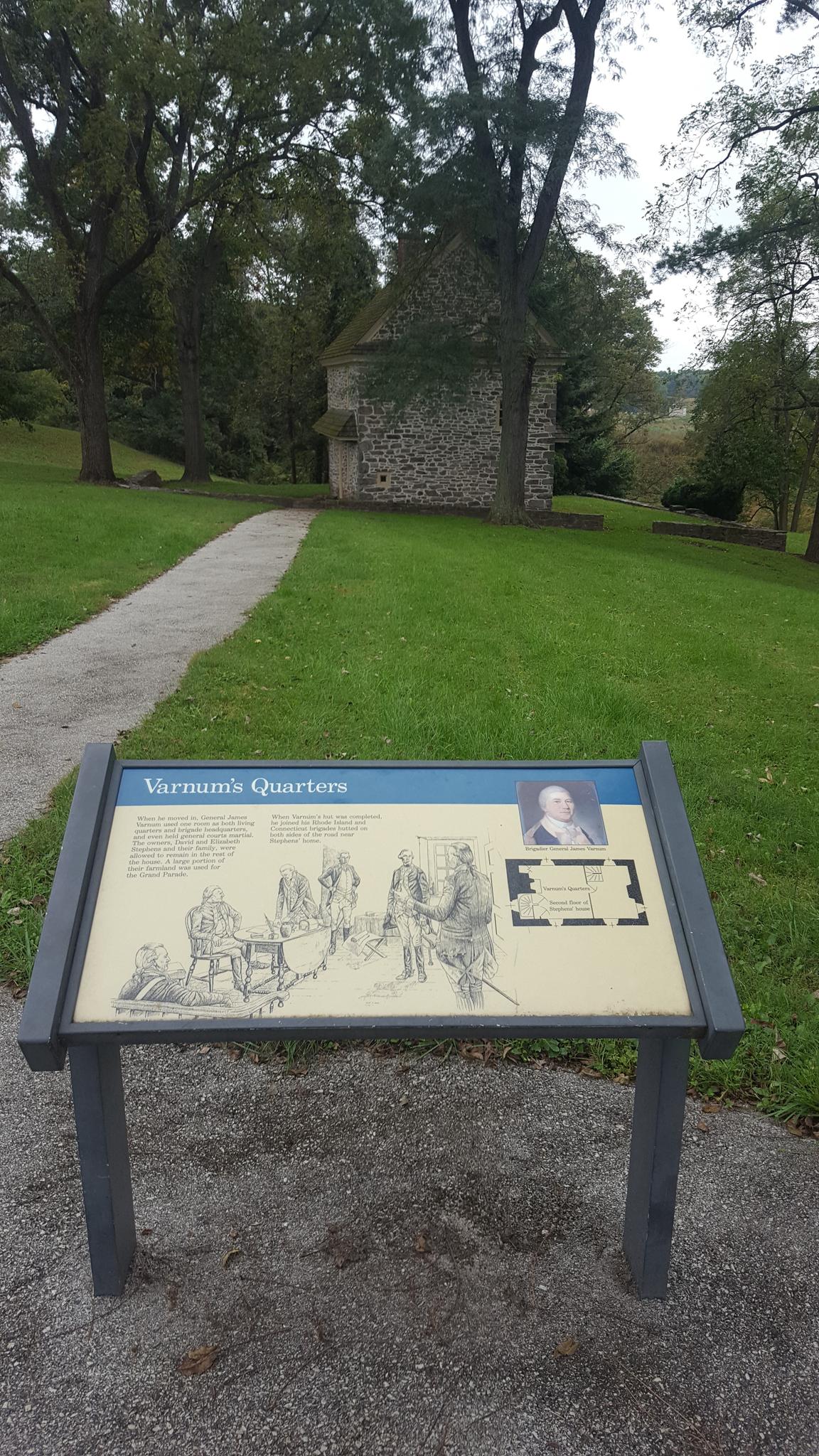 Informational sign in foreground, stone building and trees in background, overcast.