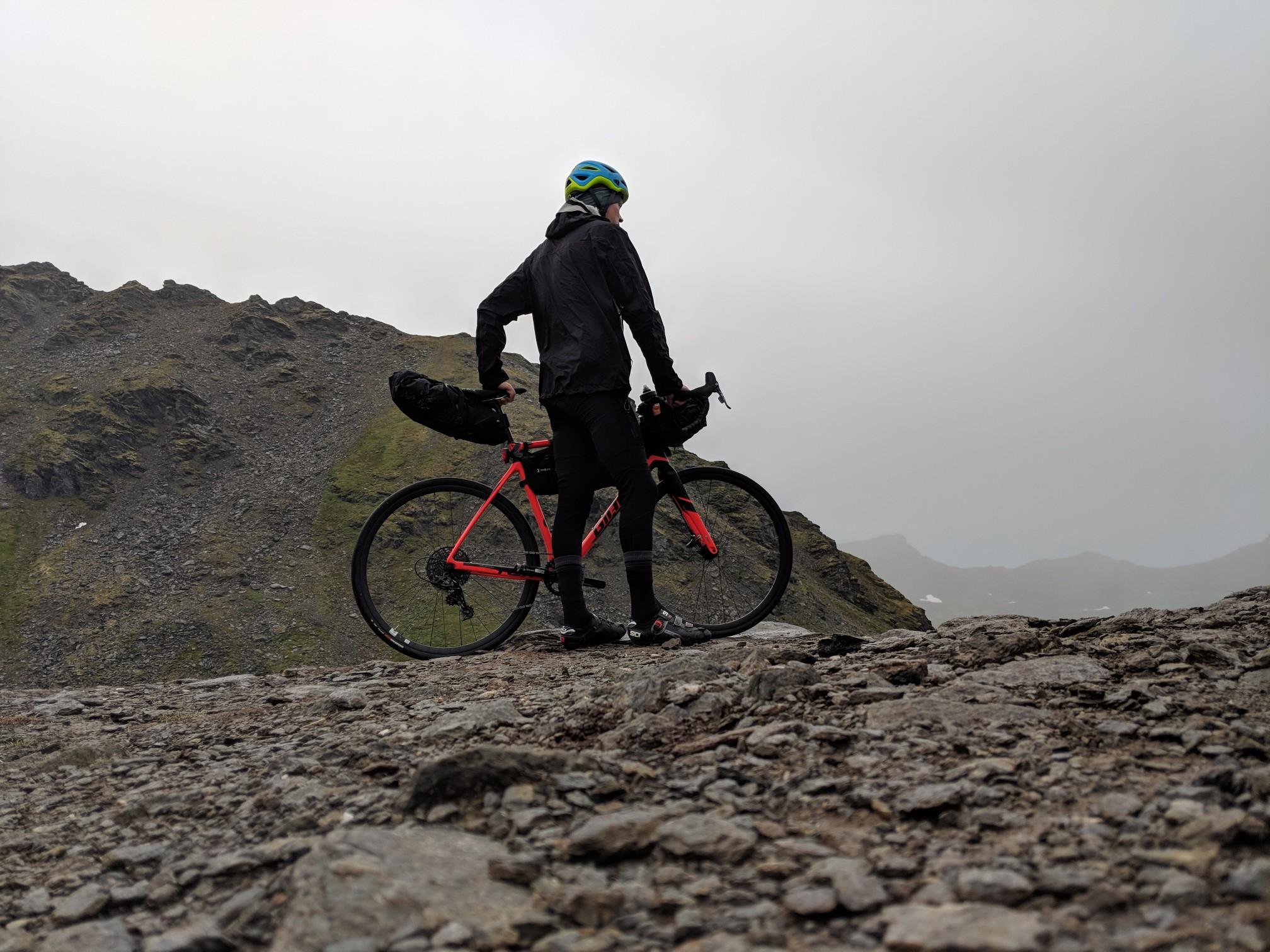 Cyclist stands with bike on rocky mountain, looking at foggy landscape.