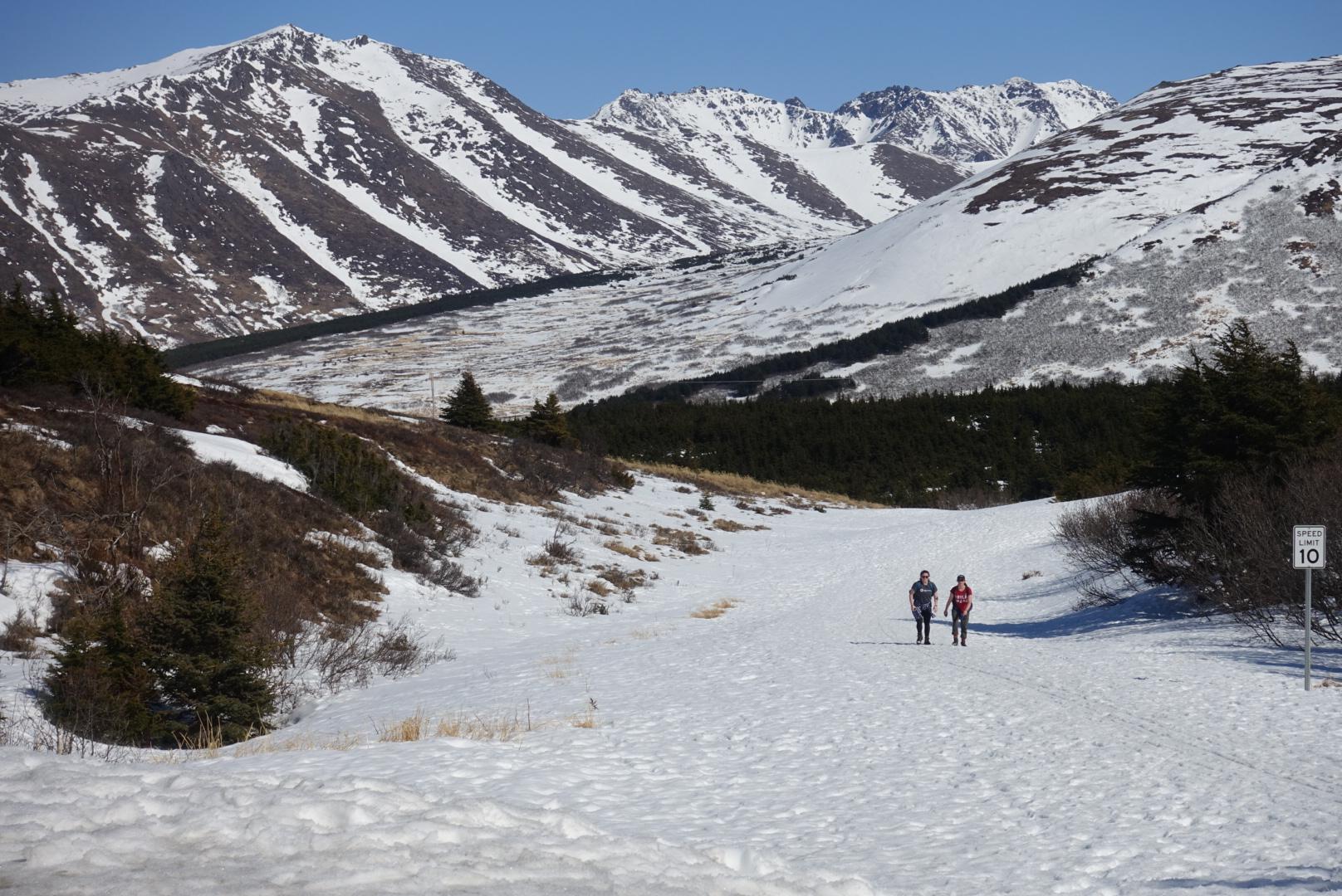 Two hikers walk on a snowy path through a valley with snow-capped mountains.