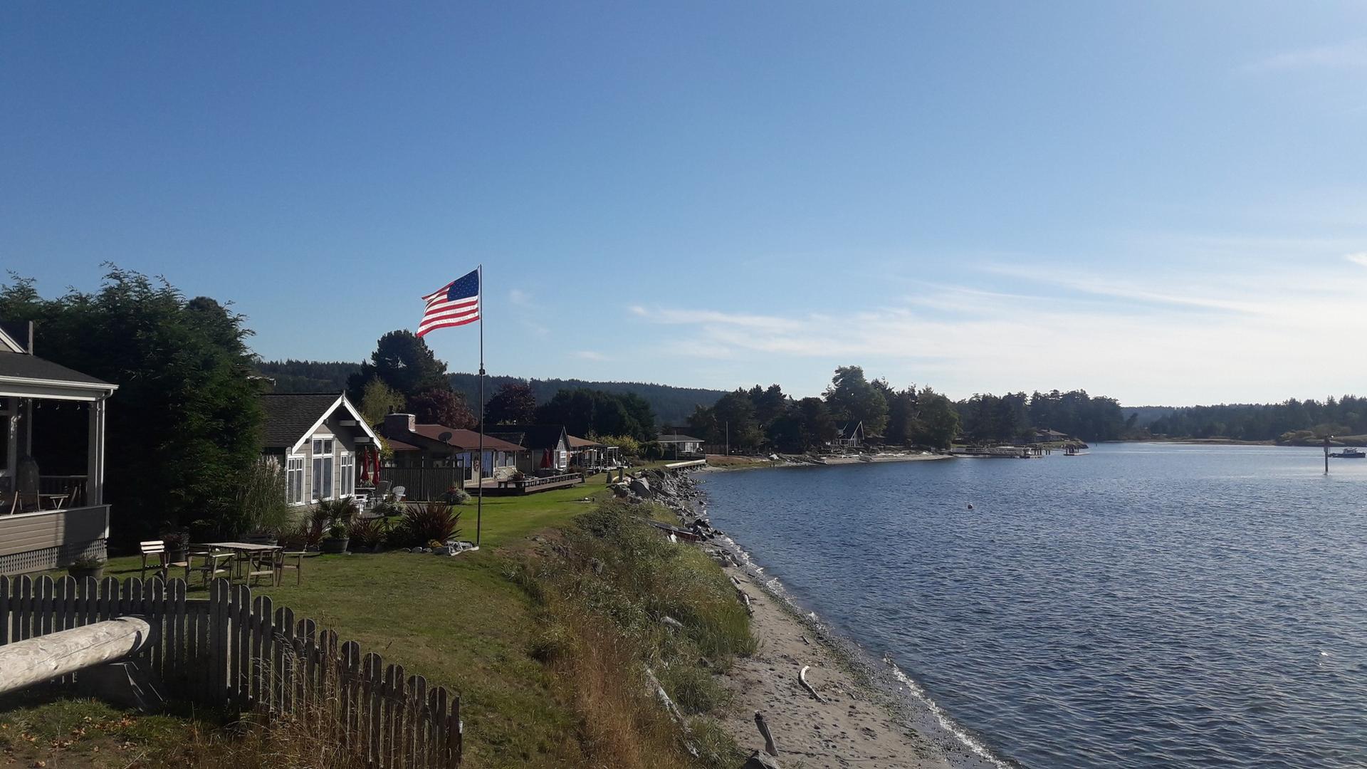 Houses and an American flag line a sunny, calm waterfront.
