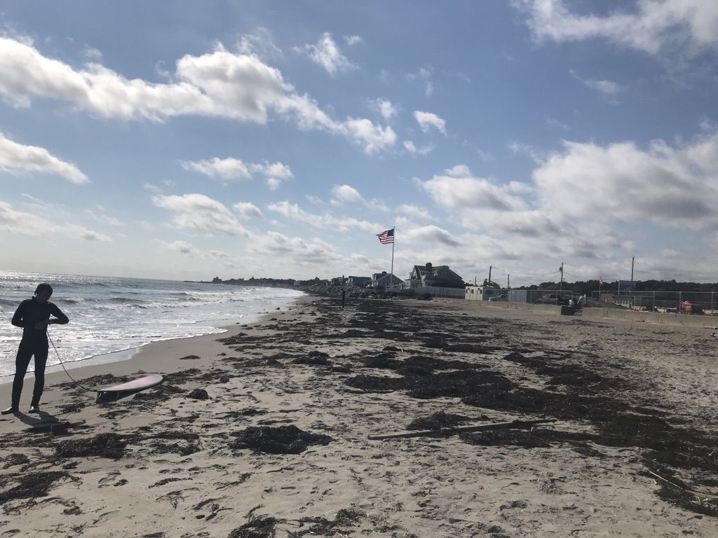 A person in a wetsuit stands on a sandy beach with waves. - East Coast Greenway: New Hampshire