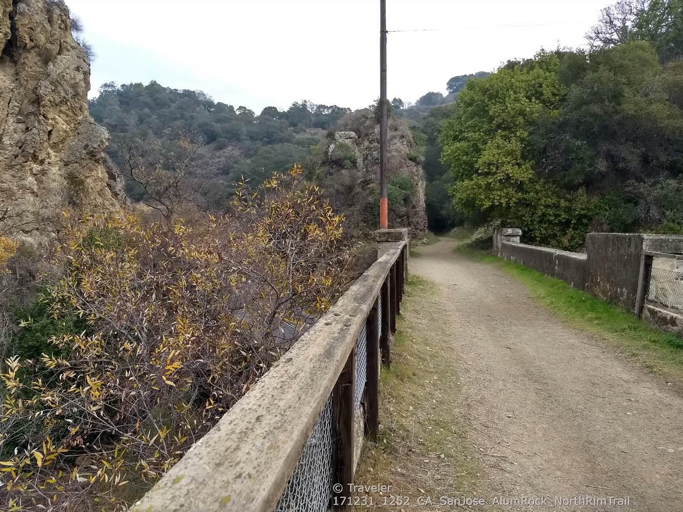 A dirt path runs beside a concrete fence, with rocky cliffs and trees.