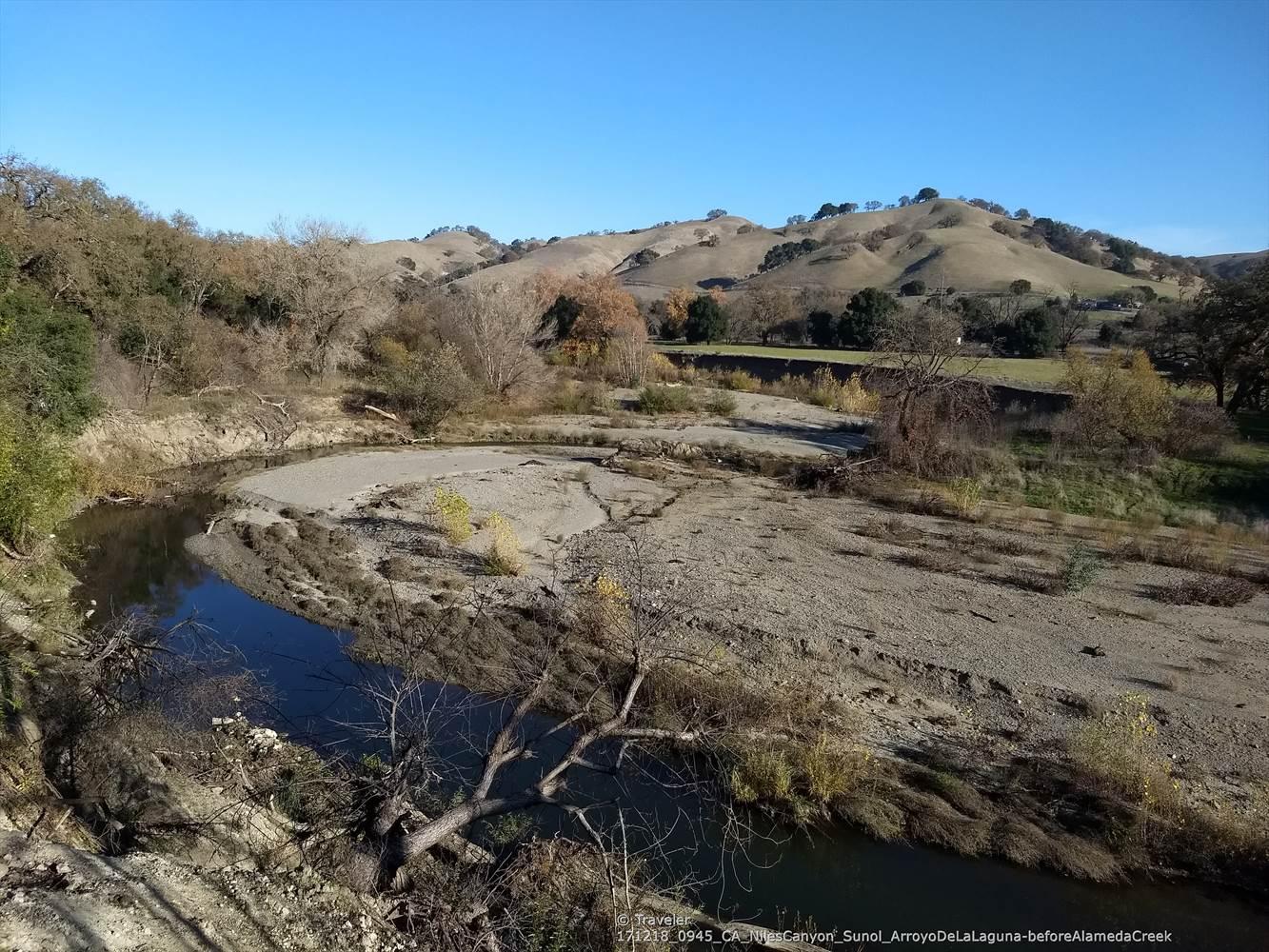 A winding creek flows through a dry, rocky riverbed with hills.