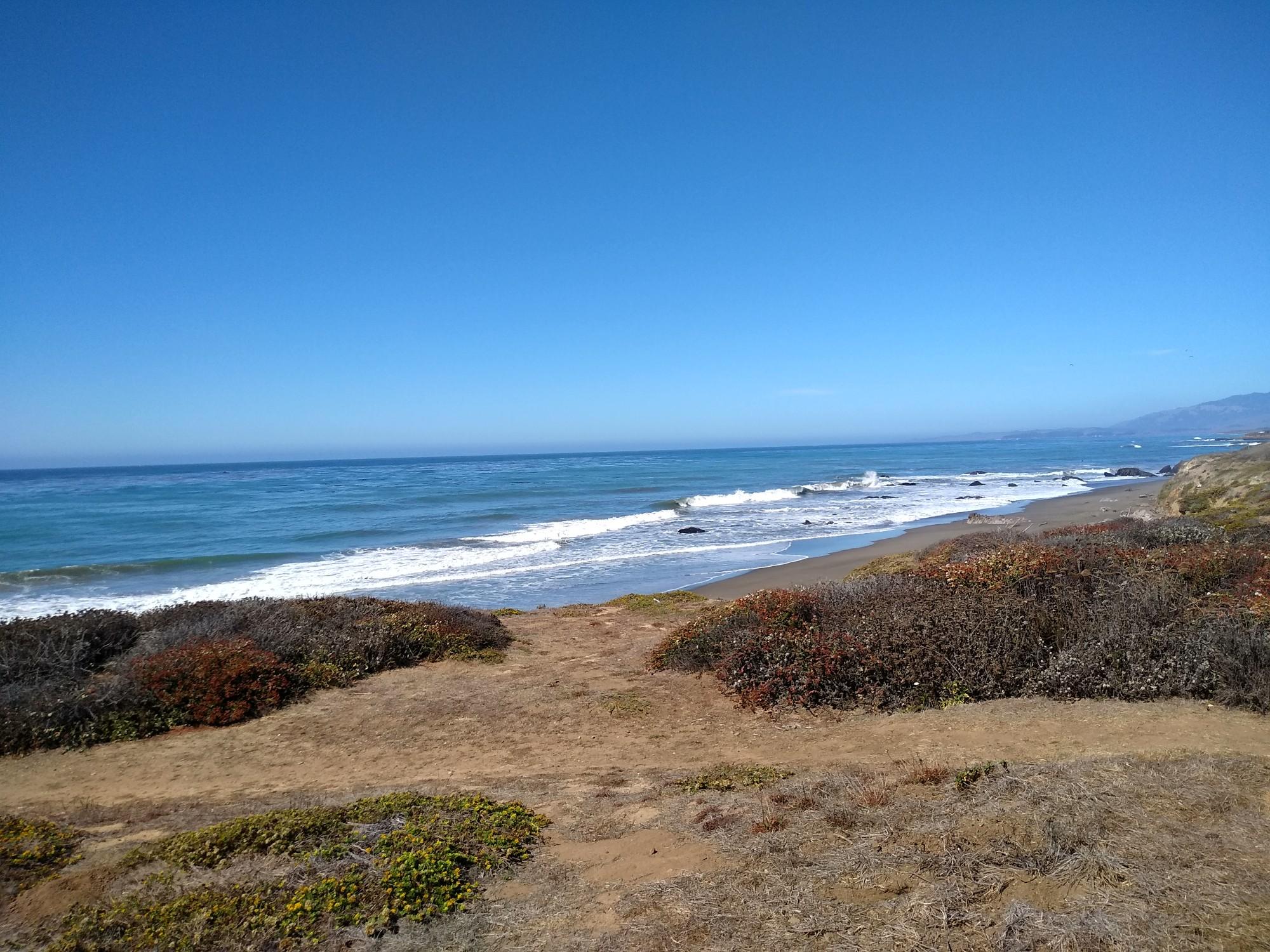 Sunny beach with waves, coastal scrub, and distant hills.