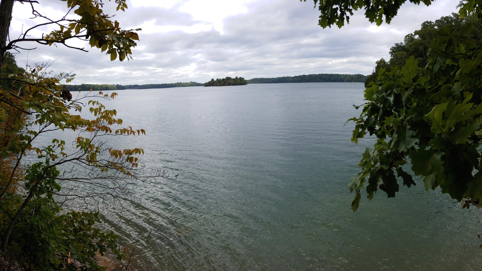 A wide, calm lake under an overcast sky, framed by green trees.