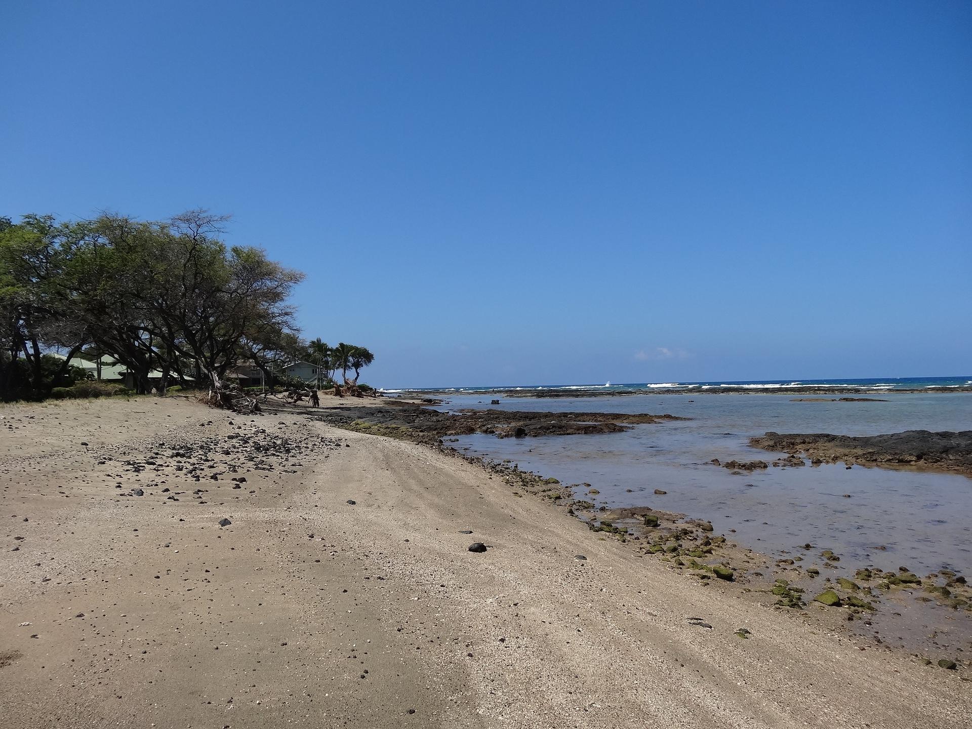 Wide sandy beach with rocky tide pools and ocean waves under clear blue sky.