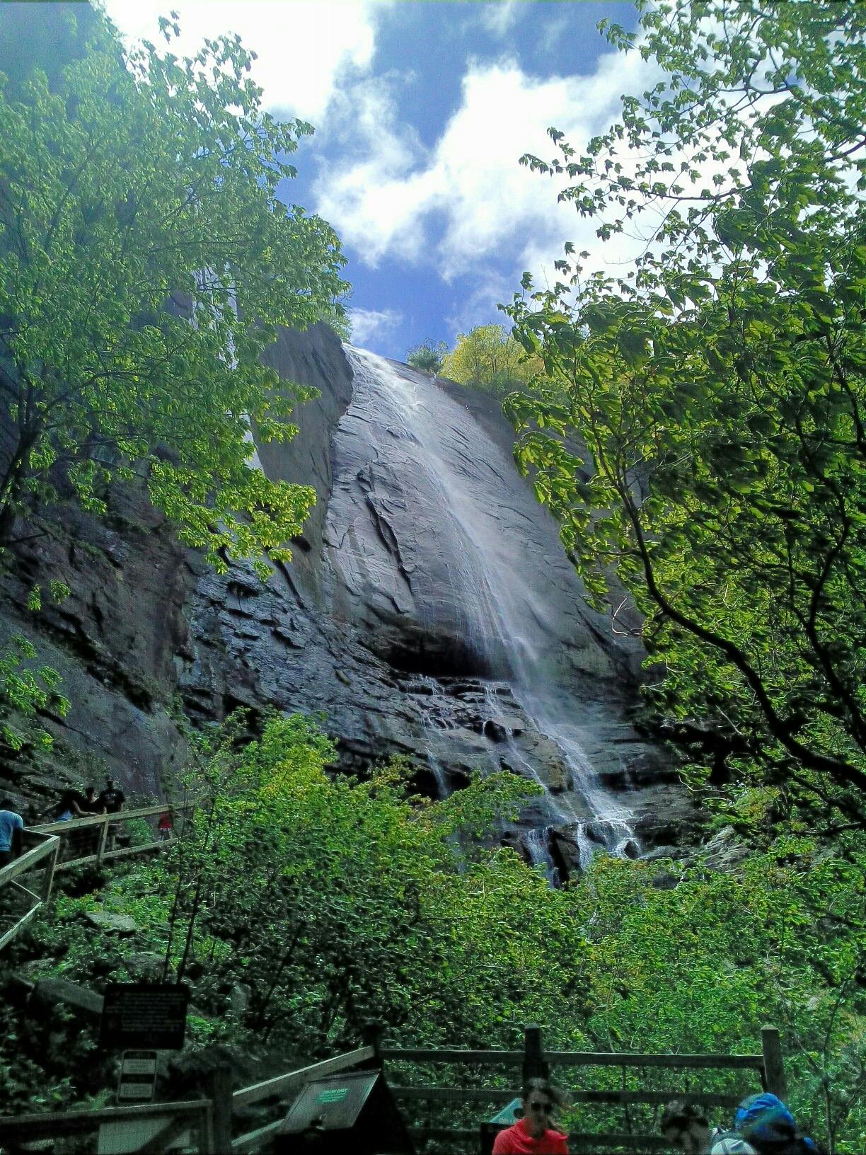 Tall waterfall cascading down a rocky cliff face, surrounded by green trees. Bright daylight, people below.