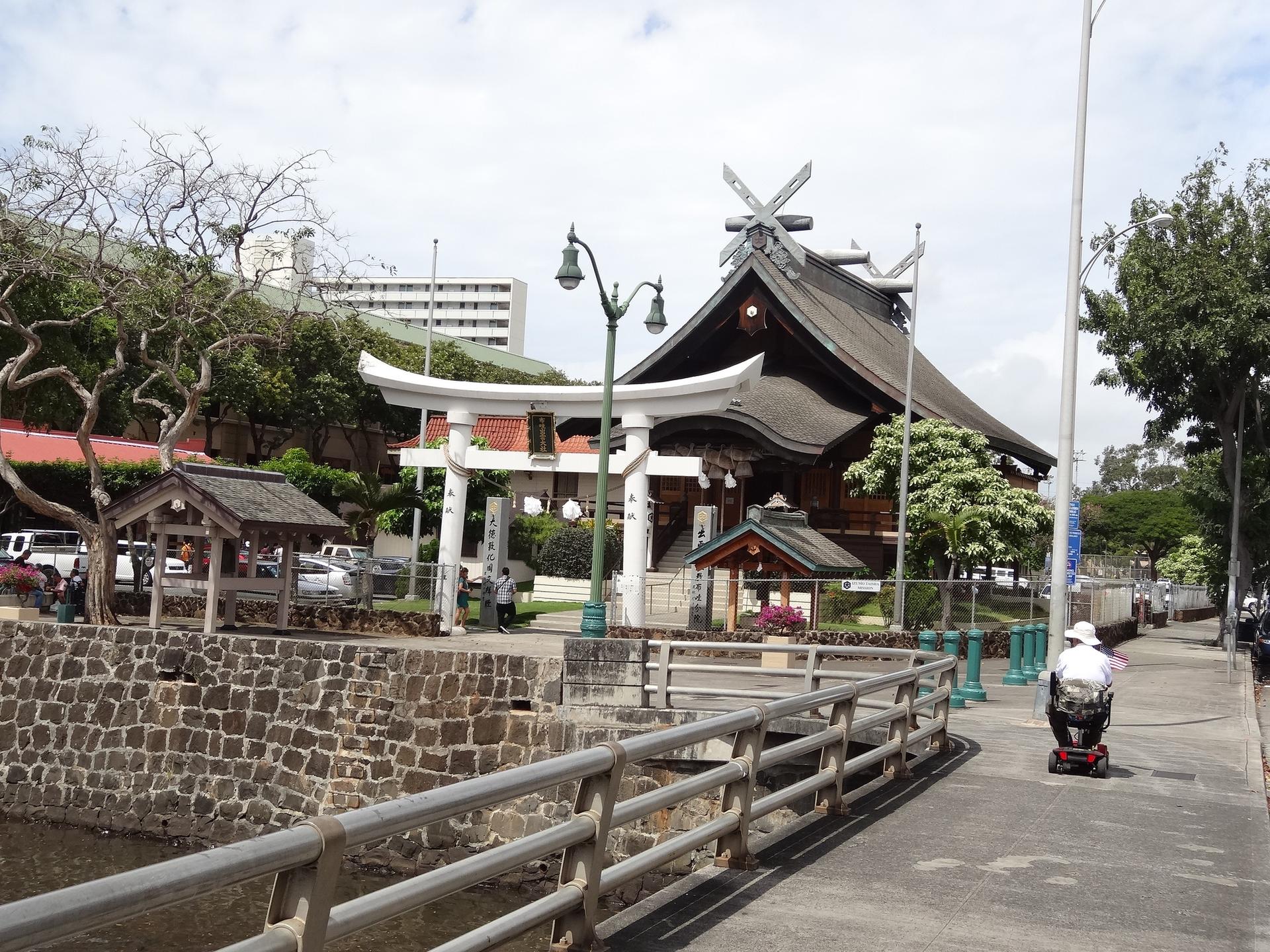 Traditional building and torii gate under overcast sky, person on scooter.