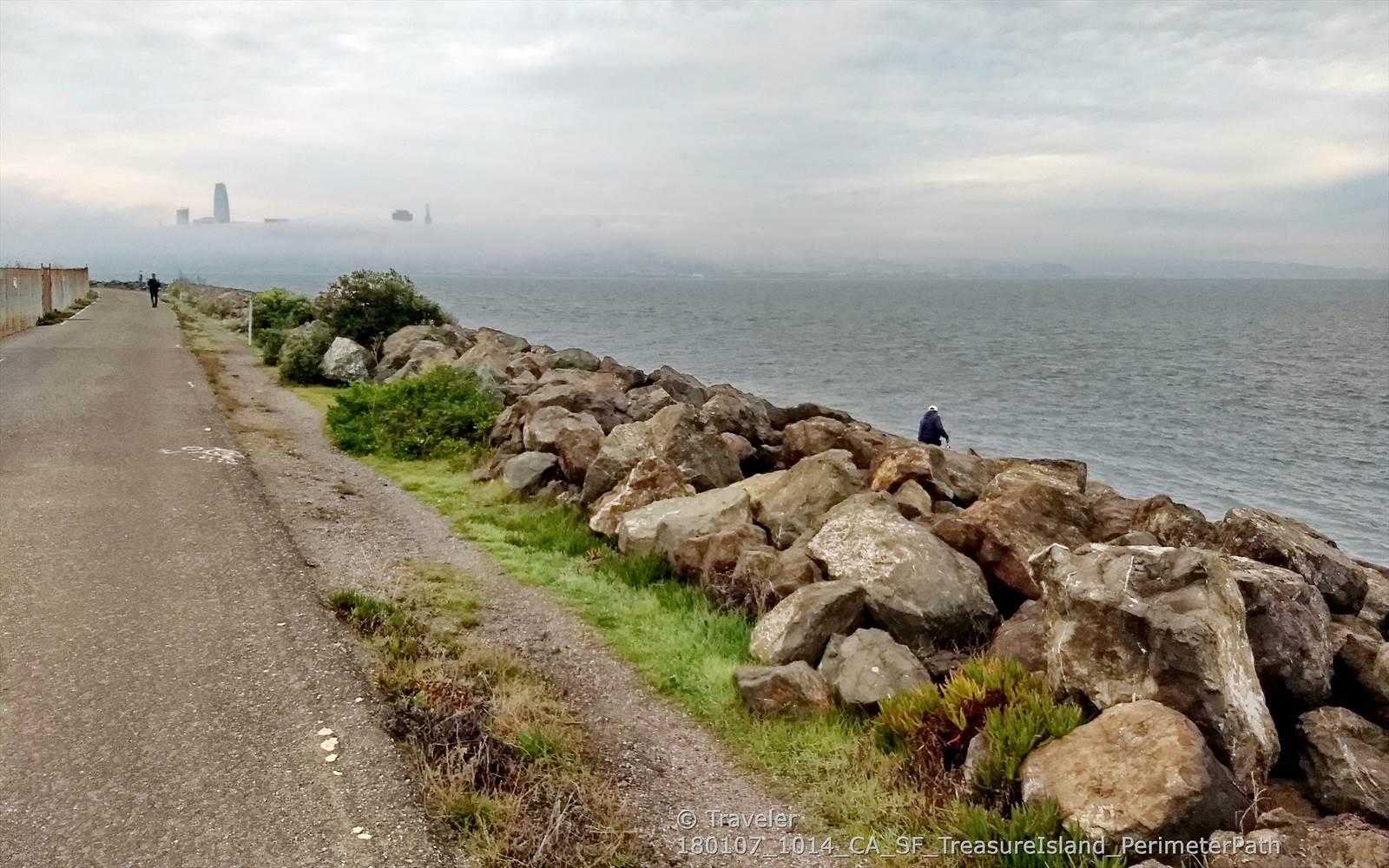Rocky shoreline next to a path, with a person fishing under an overcast sky.