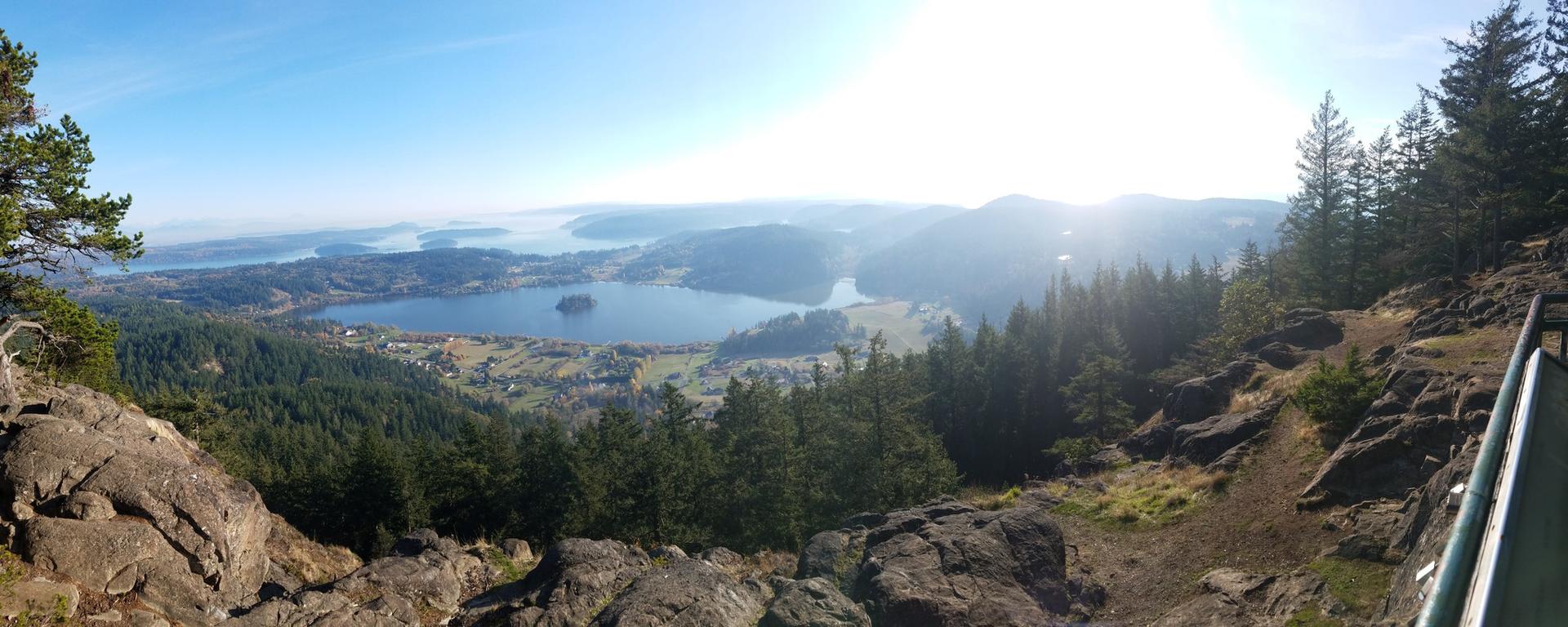 Panoramic view from rocky cliffs overlooking a lake and distant water, bright sun.