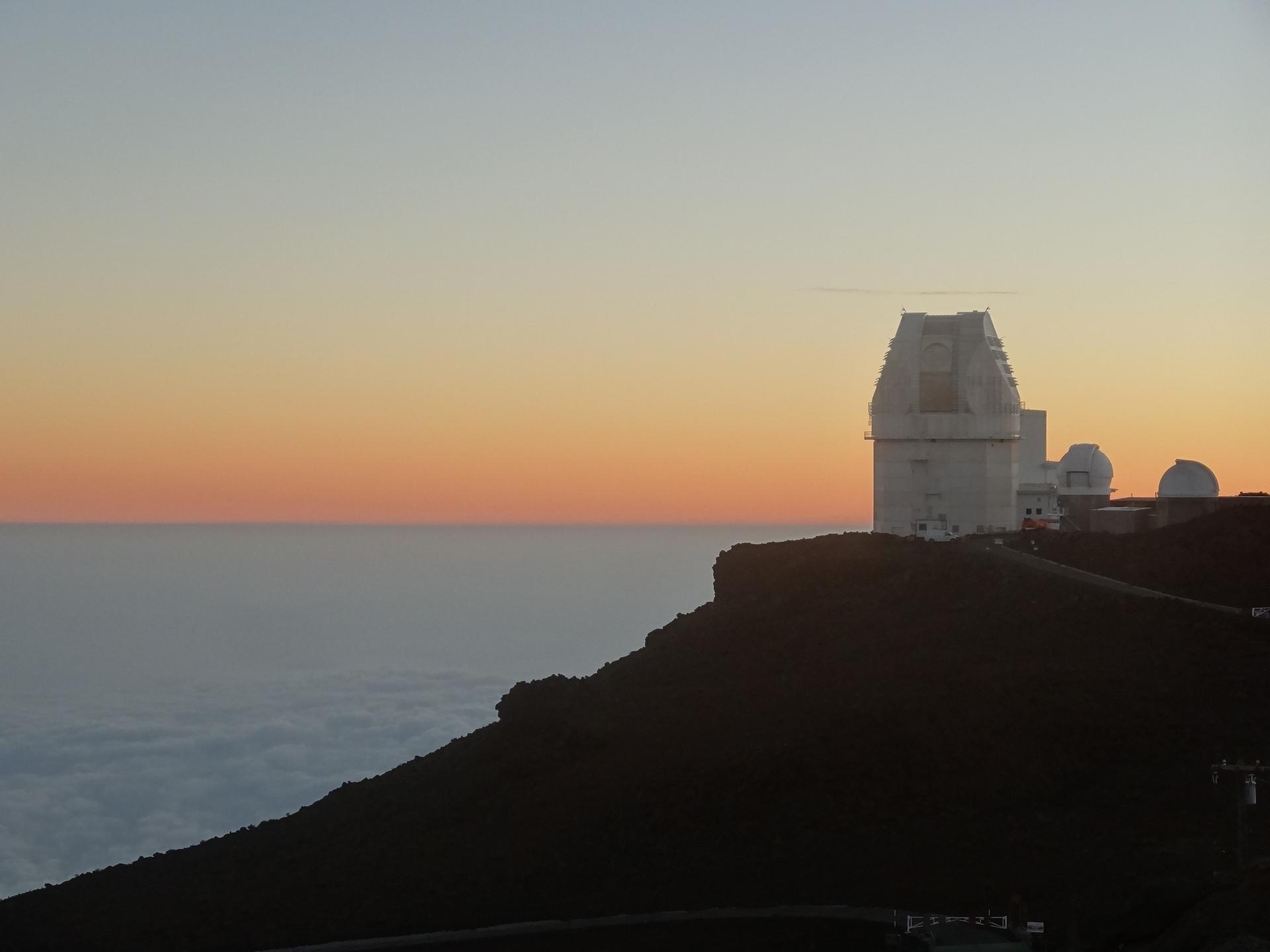White observatory domes silhouetted against a colorful sunset sky above clouds.