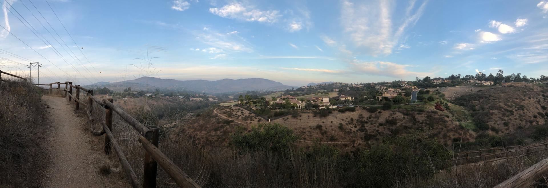 A dirt trail with a wooden fence overlooks a vast, dry, hilly landscape.