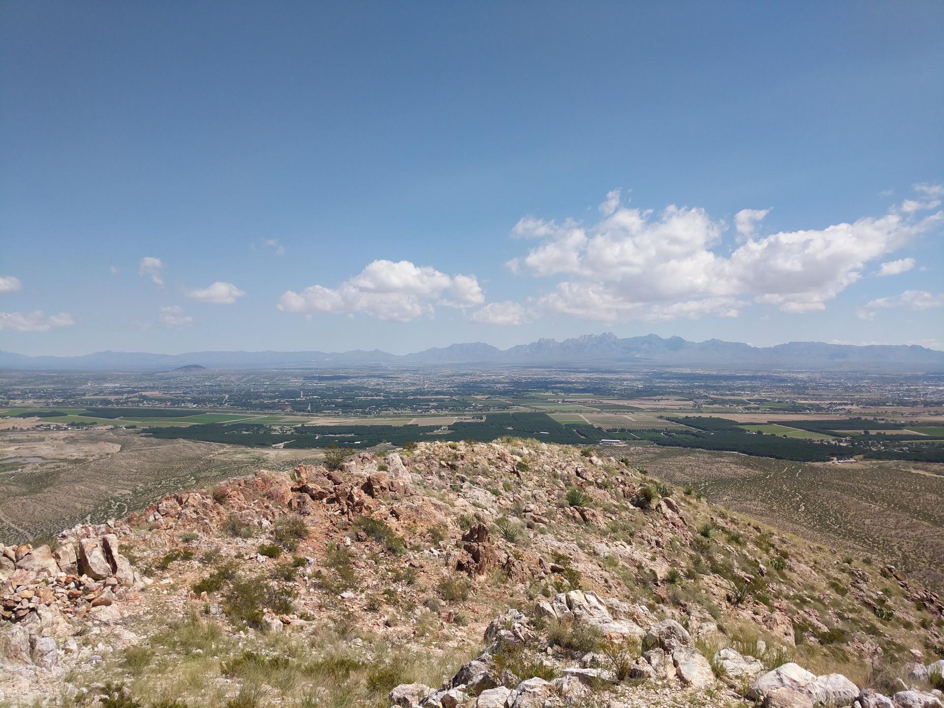 Rocky hilltop overlooking a vast valley and distant mountains under a bright sky.