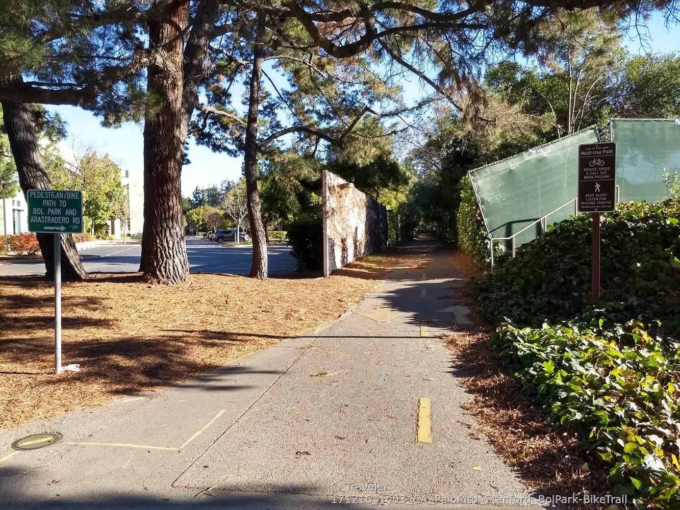 A paved path winds through a shaded area with large trees and signs.