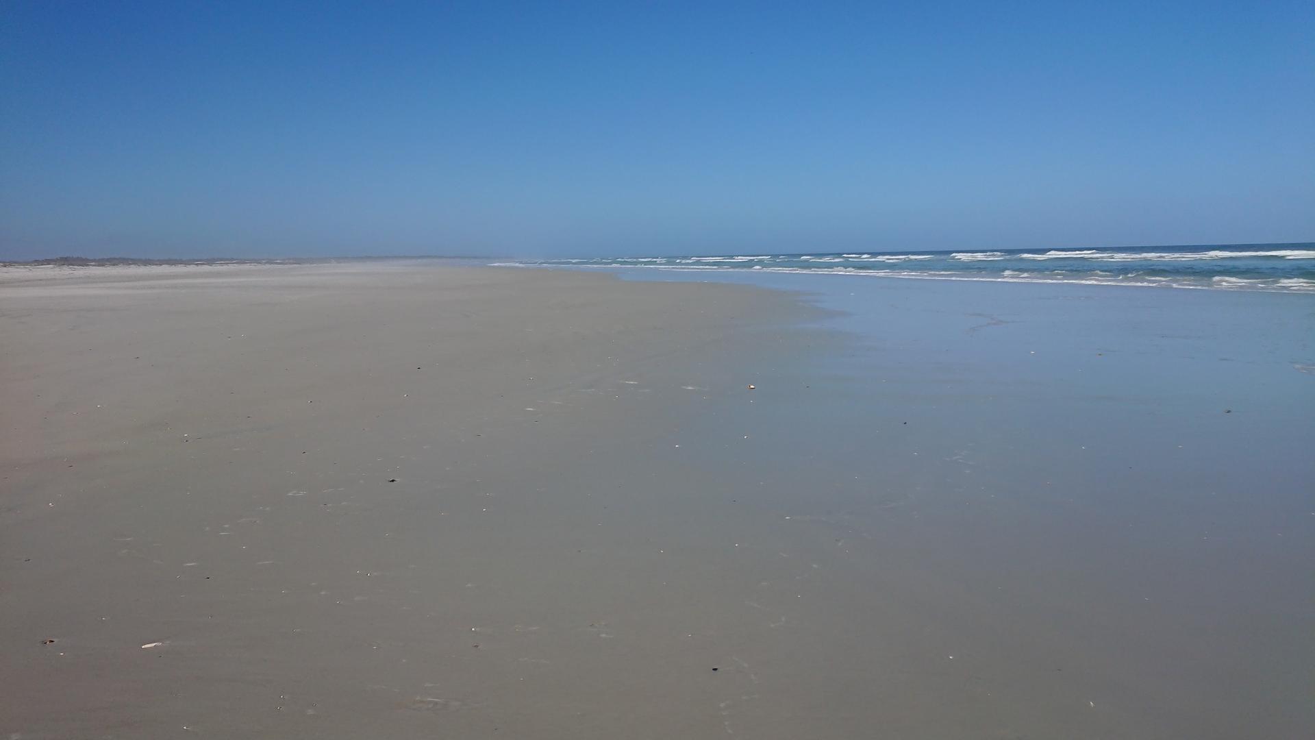 A wide, empty sandy beach with gentle waves under a clear blue sky.
