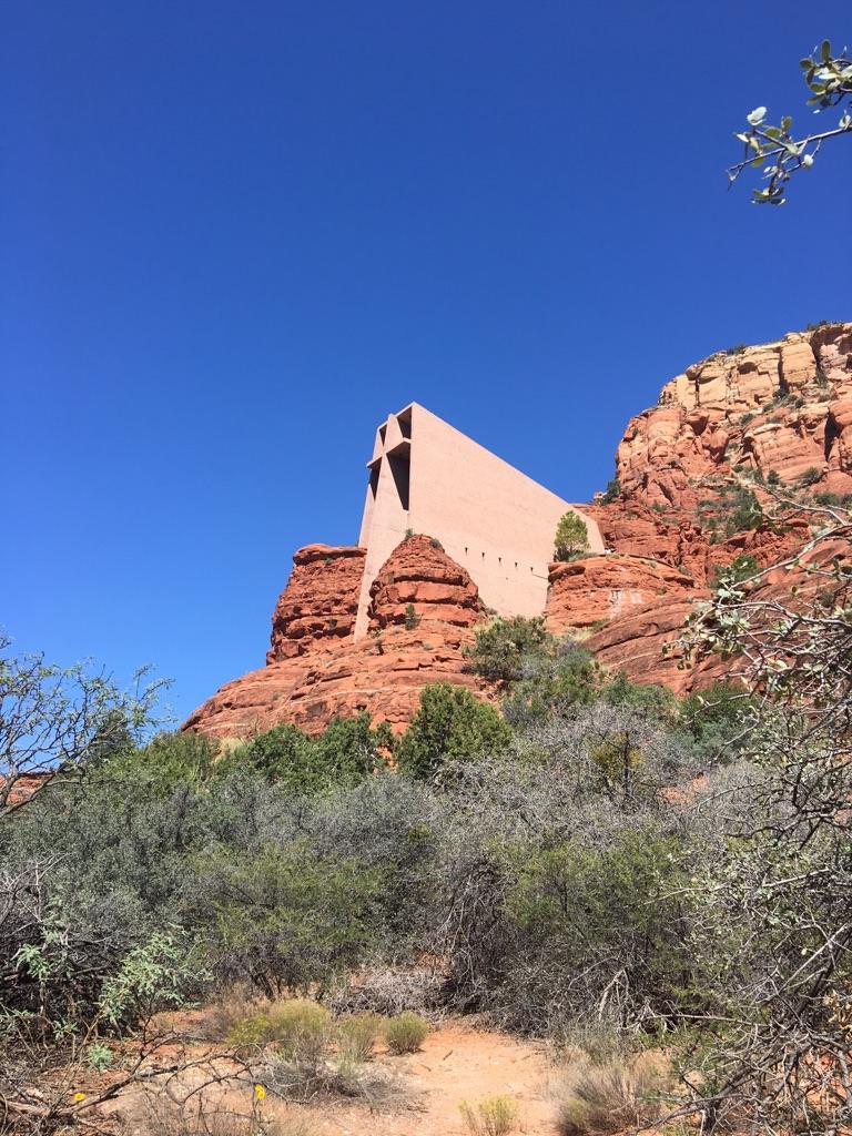 Modern chapel built into a red rock cliff, surrounded by green shrubs.
