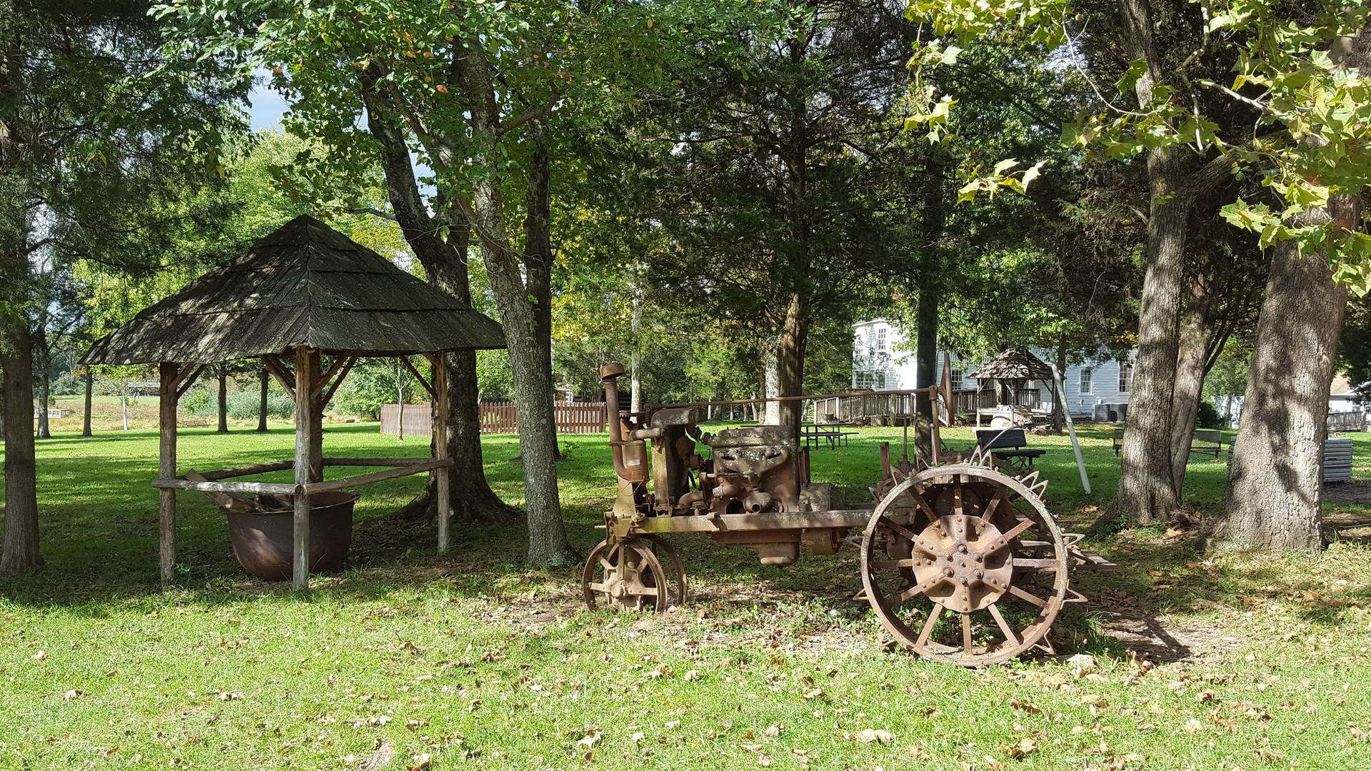 Rustic wooden shelter and old farm equipment under trees in dappled sunlight.