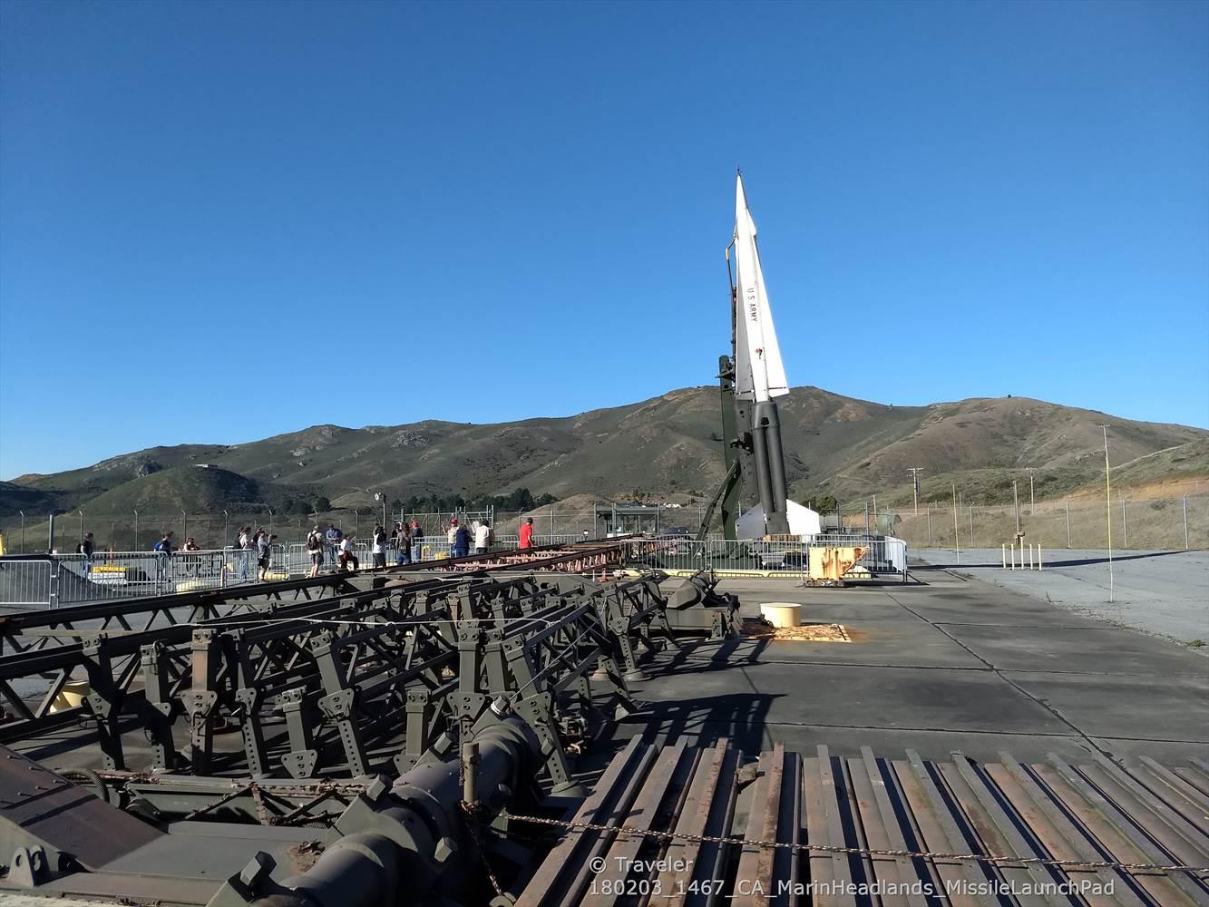 People observing a missile on a launchpad with hills in the background.