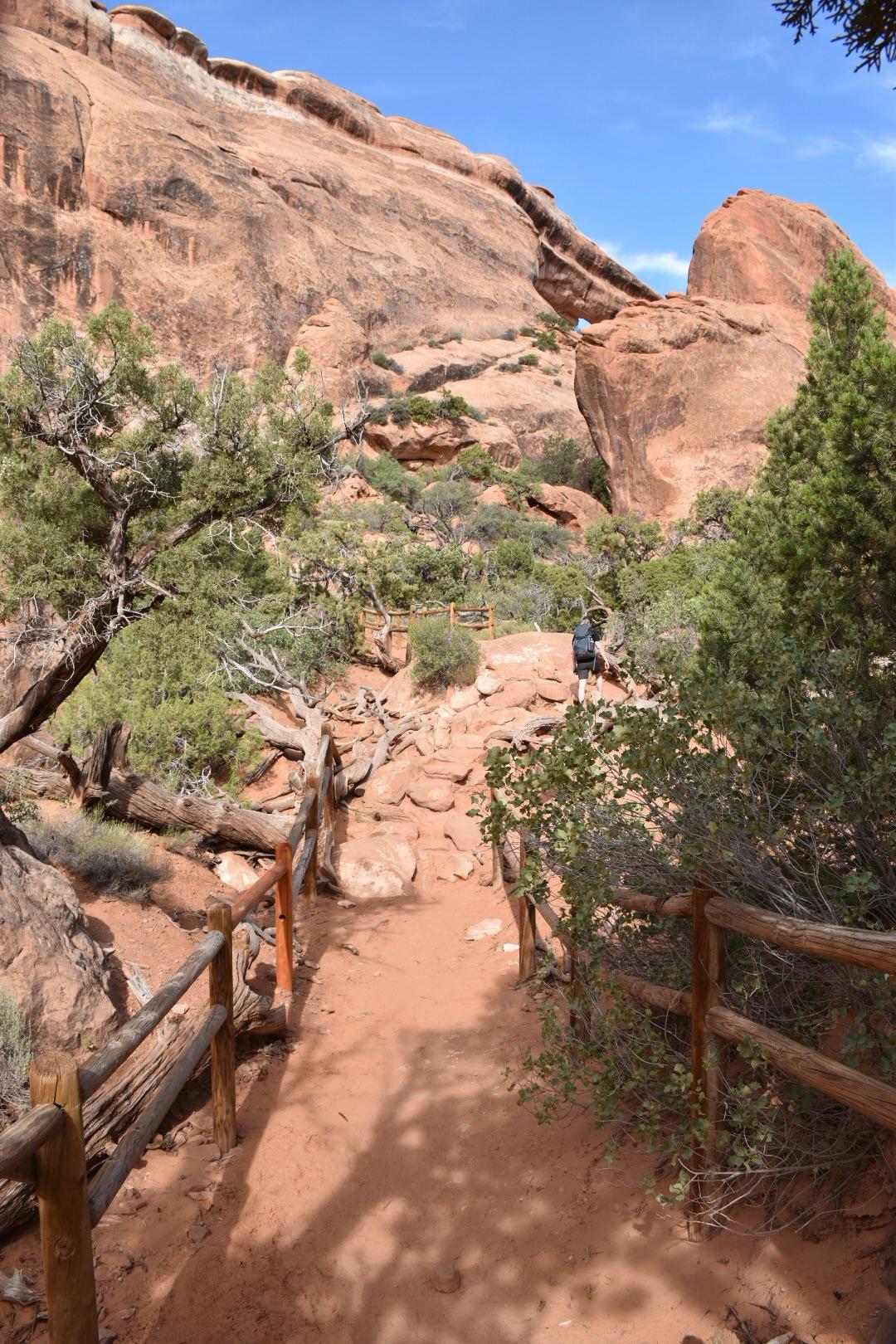 A person walks up a rocky path with wooden fences and red rock walls.