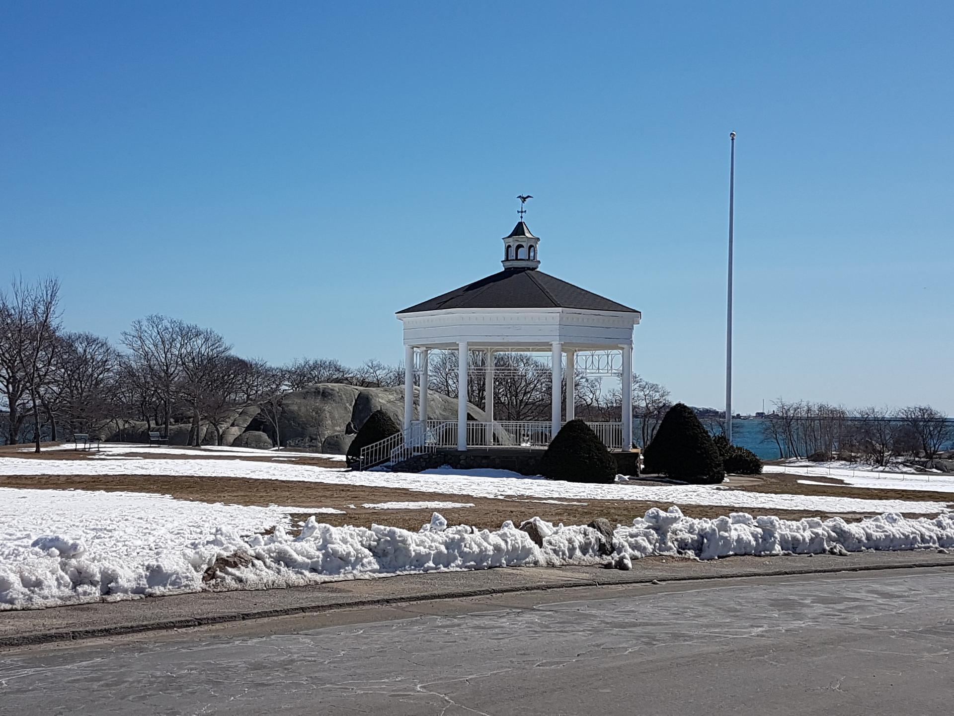 White gazebo in a snowy park with bare trees under a clear blue sky.