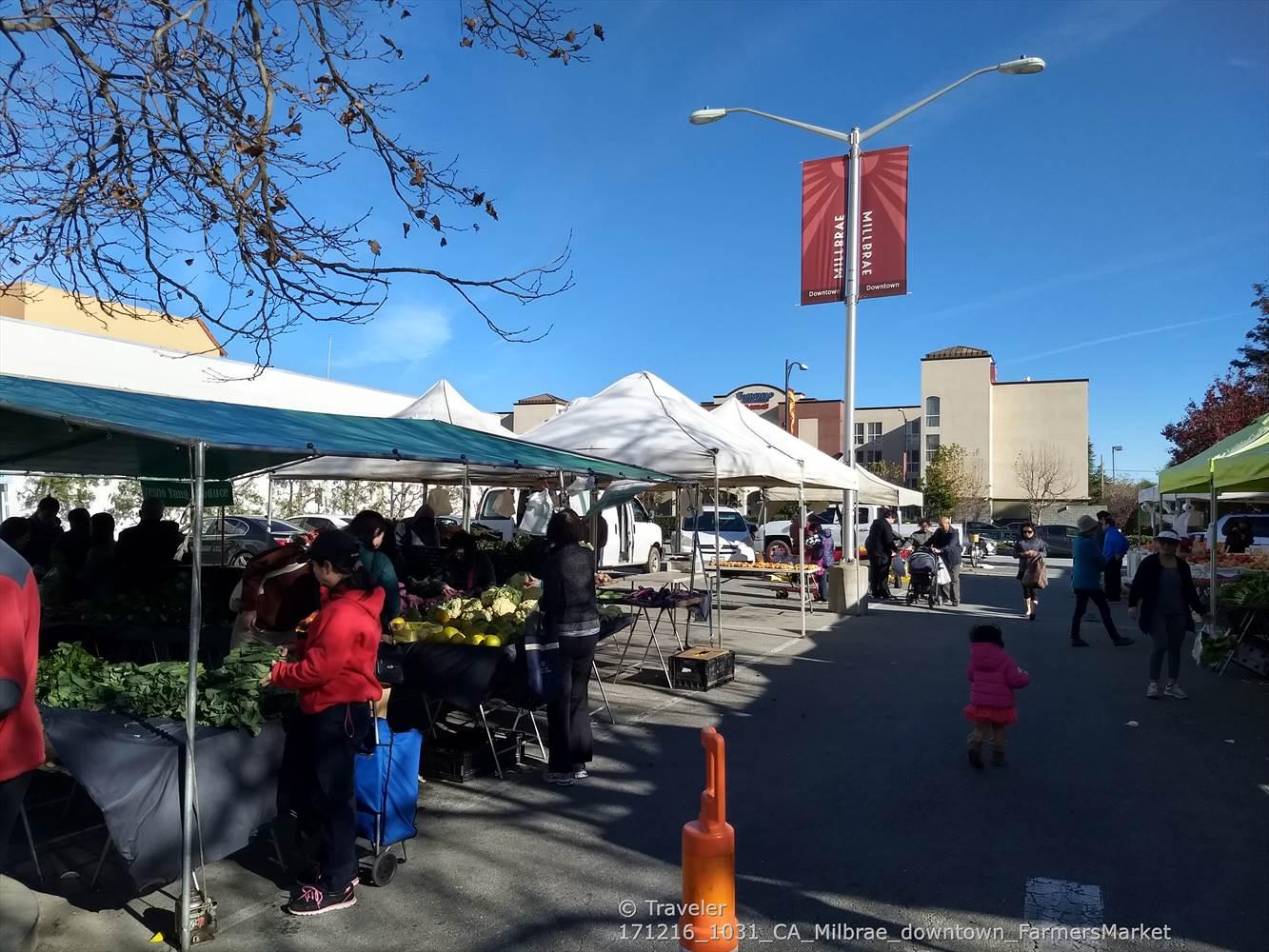 Bustling outdoor market with white tents, people shopping, under a bright blue sky.