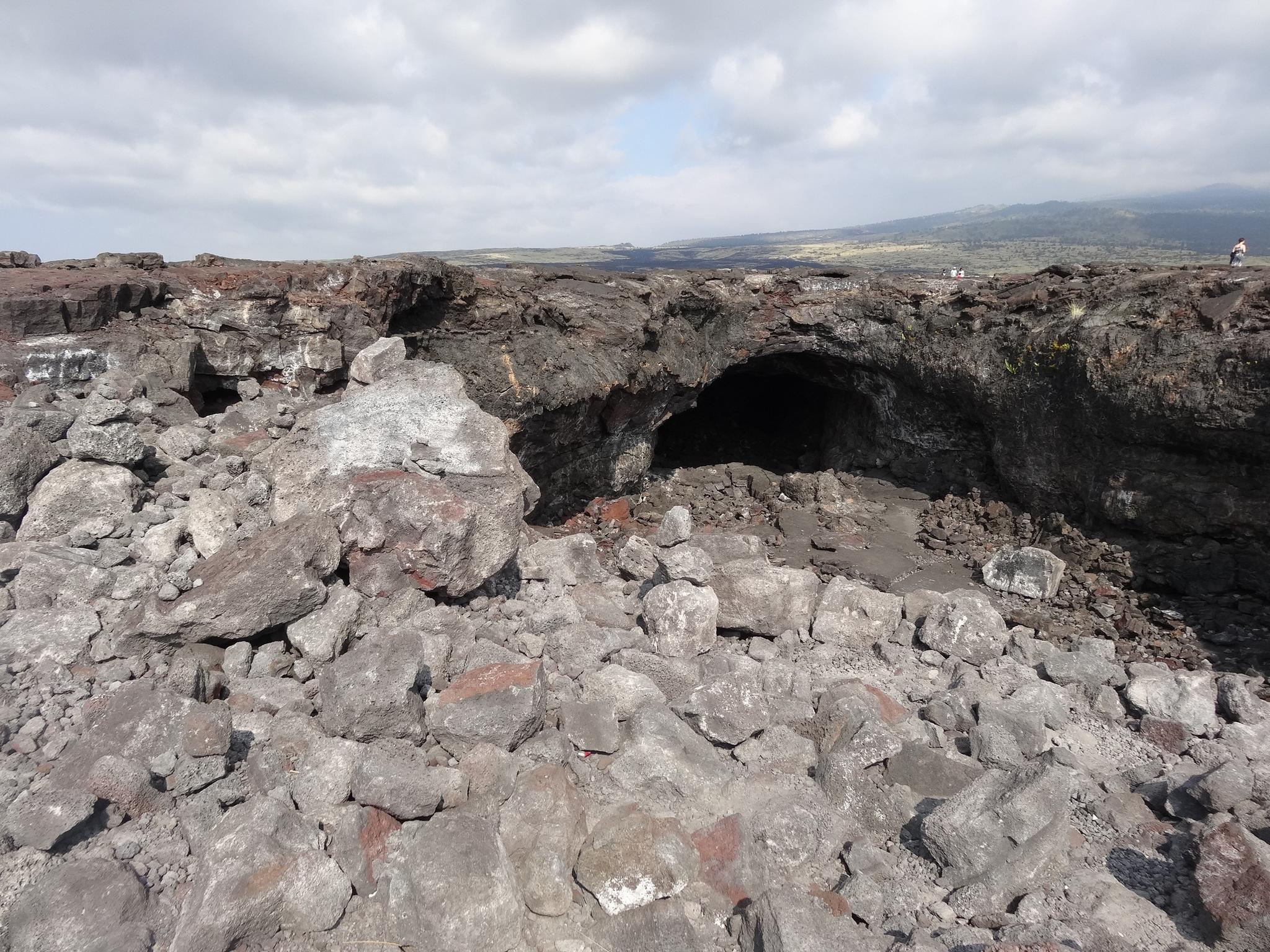Dark cave entrance surrounded by jagged volcanic rocks under a cloudy sky.