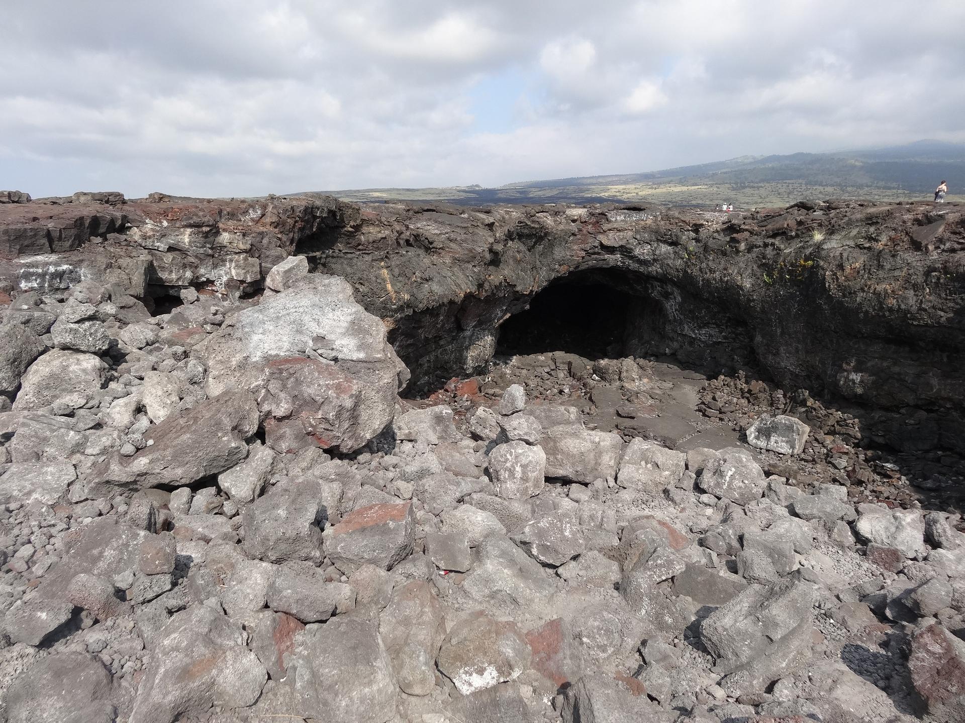 Dark cave entrance surrounded by jagged volcanic rocks under a cloudy sky.