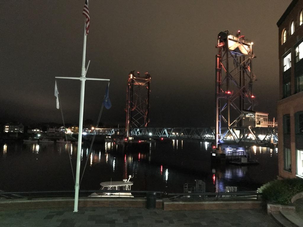 Nighttime view of a waterfront with illuminated bridge towers, buildings, and a flagpole.