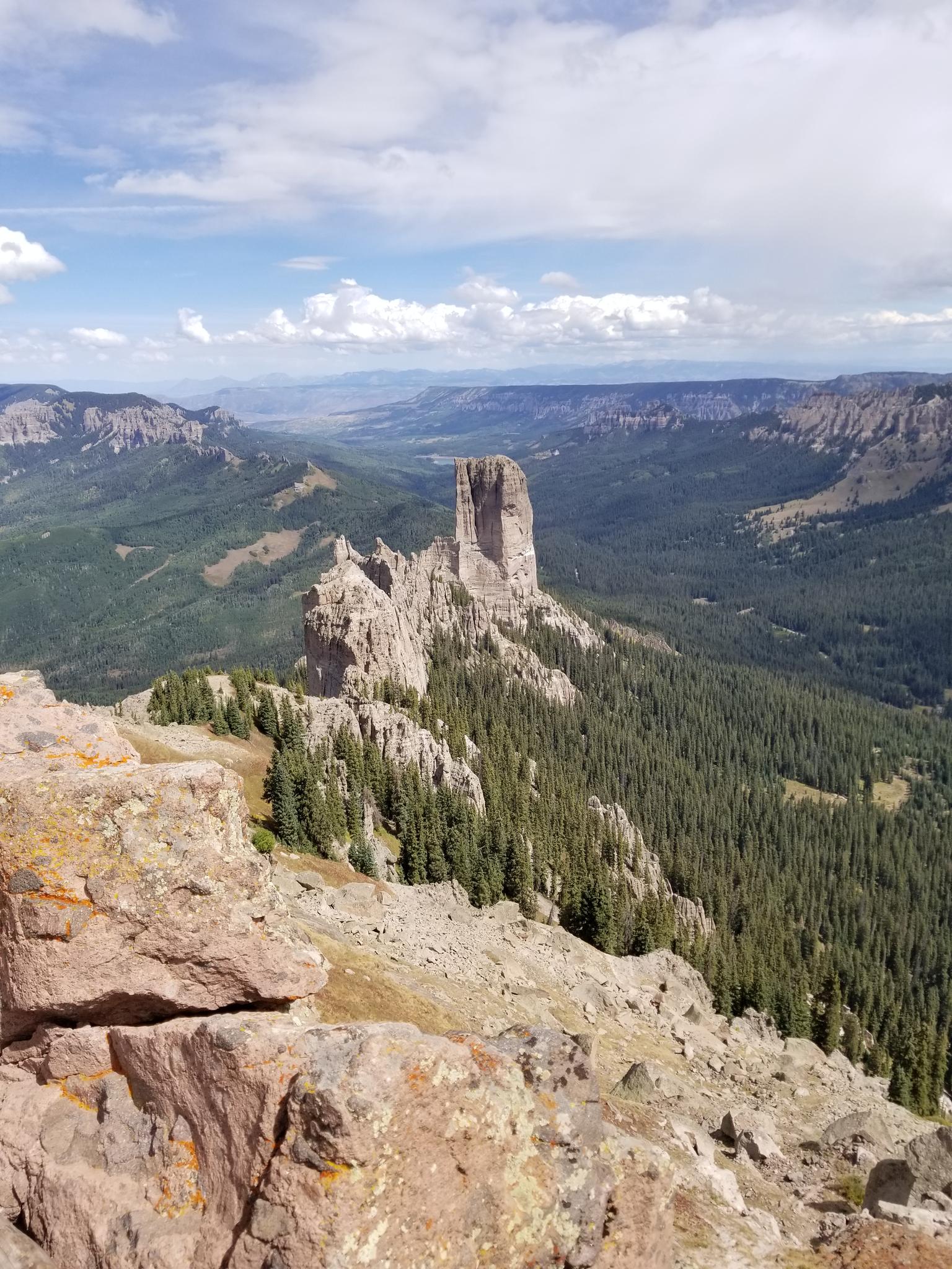 Tall rock pinnacle rises from a green forest under a partly cloudy sky.