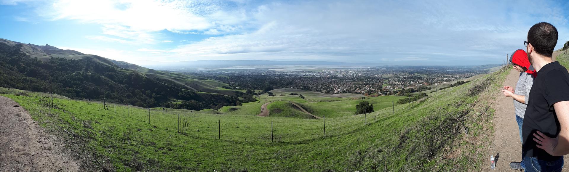 Two people admire a panoramic view of green hills and a distant city.