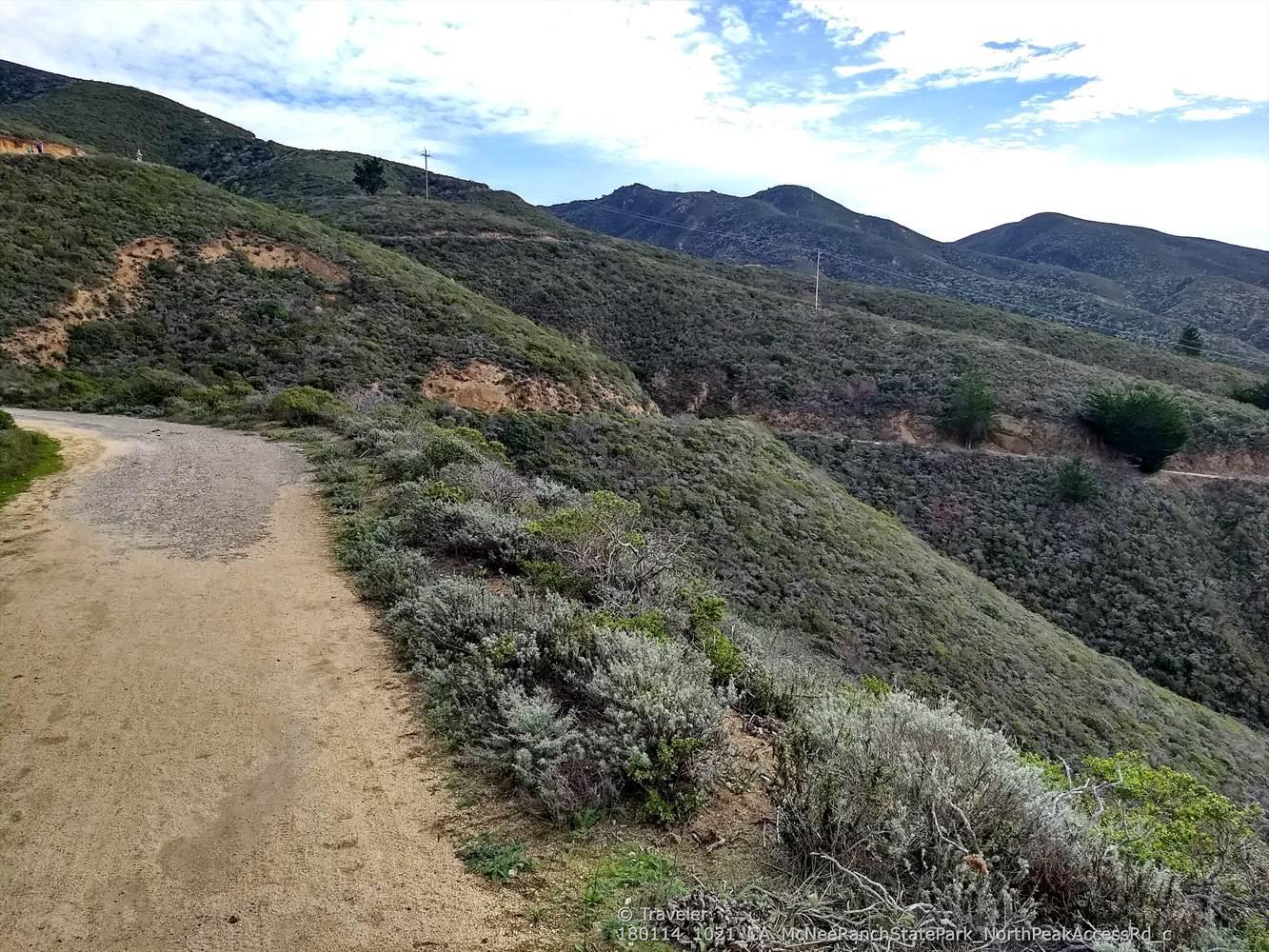 A dirt road winds up a steep, brush-covered hillside under a cloudy sky.