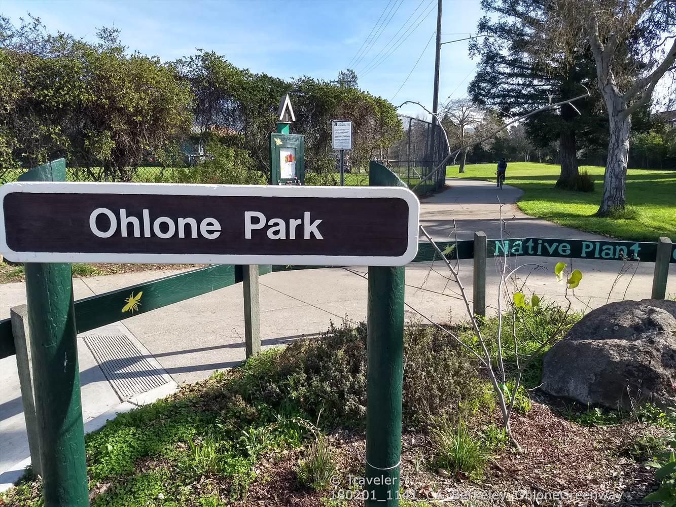 A park sign on green posts, surrounded by lush foliage and a path.