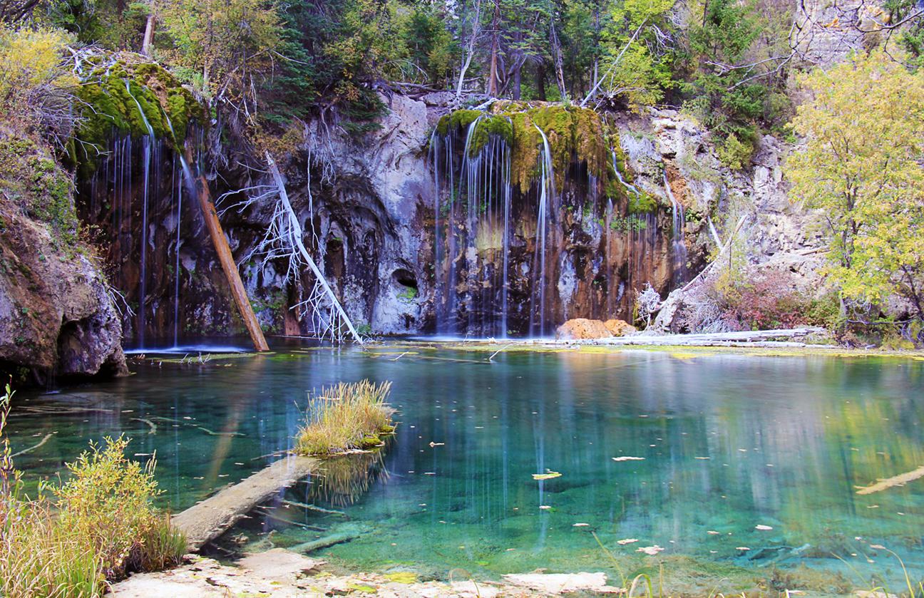 Turquoise lake with multiple waterfalls cascading over mossy rocks and fallen trees.
