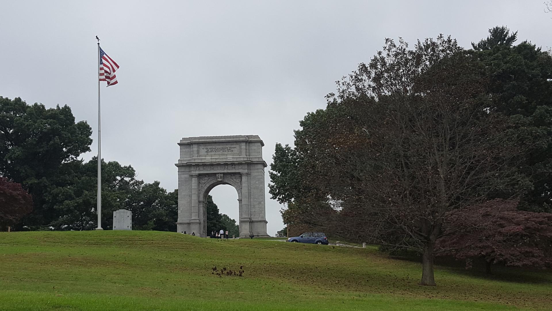 Stone arch monument on a grassy hill with an American flag under an overcast sky.