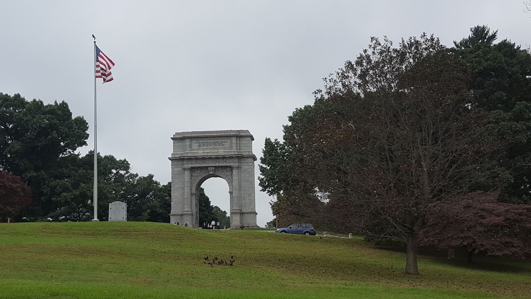 Stone arch monument on a grassy hill with an American flag under an overcast sky. - Joseph Plumb Martin Trail