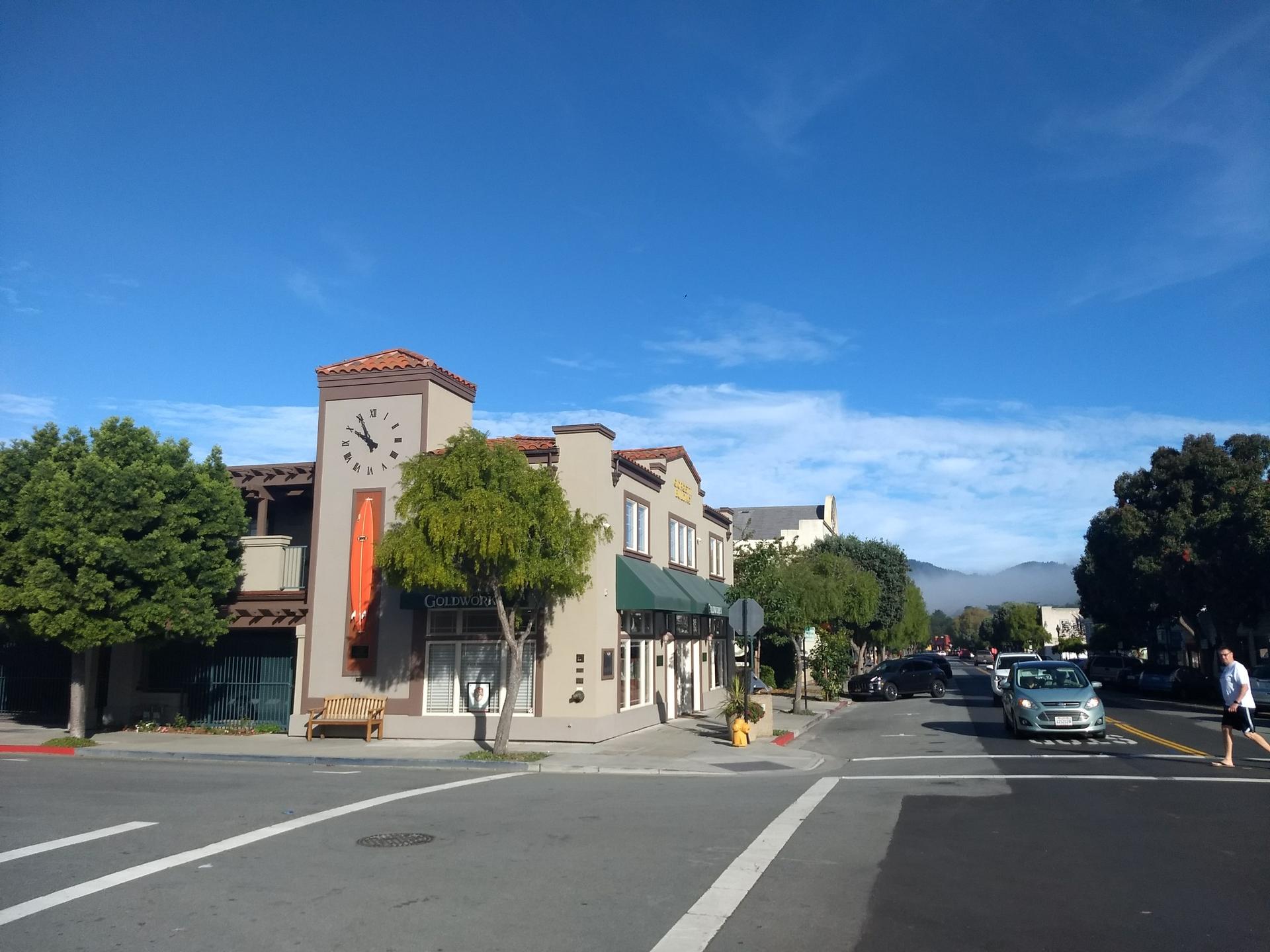 Sunny street corner with buildings, a clock tower, and a person crossing.