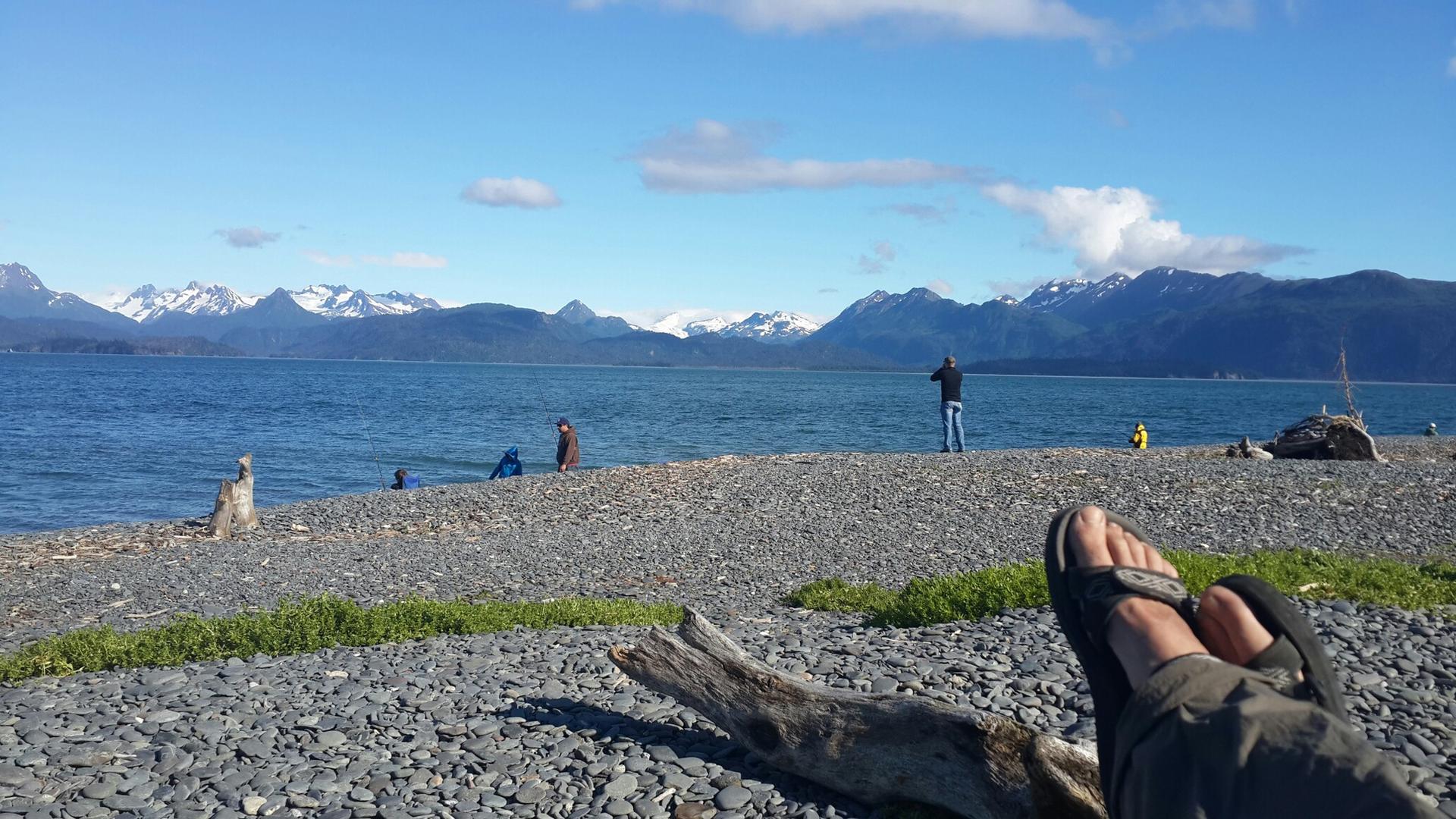 Person's feet on a rocky beach, people fishing, and snowy mountains.