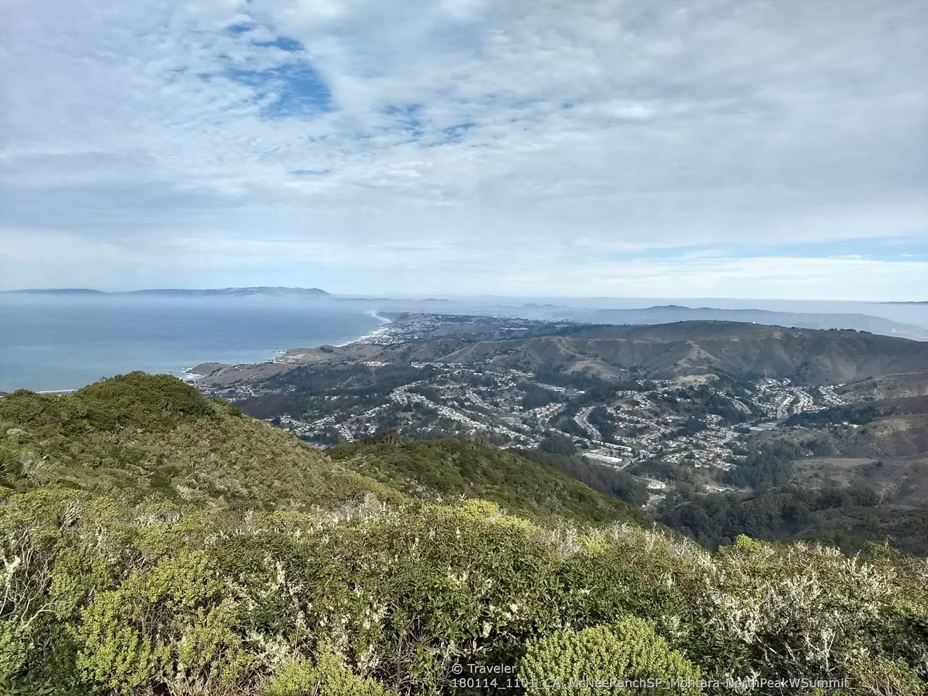 Expansive view of a coastal town, ocean, and hills under cloudy sky.