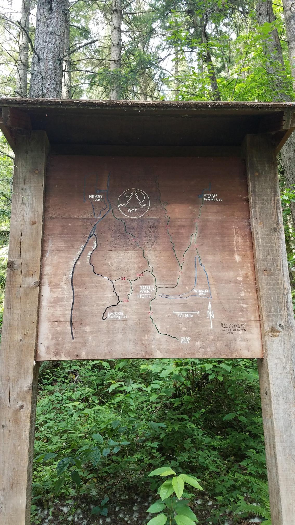 Weathered wooden trail map sign surrounded by green forest foliage.